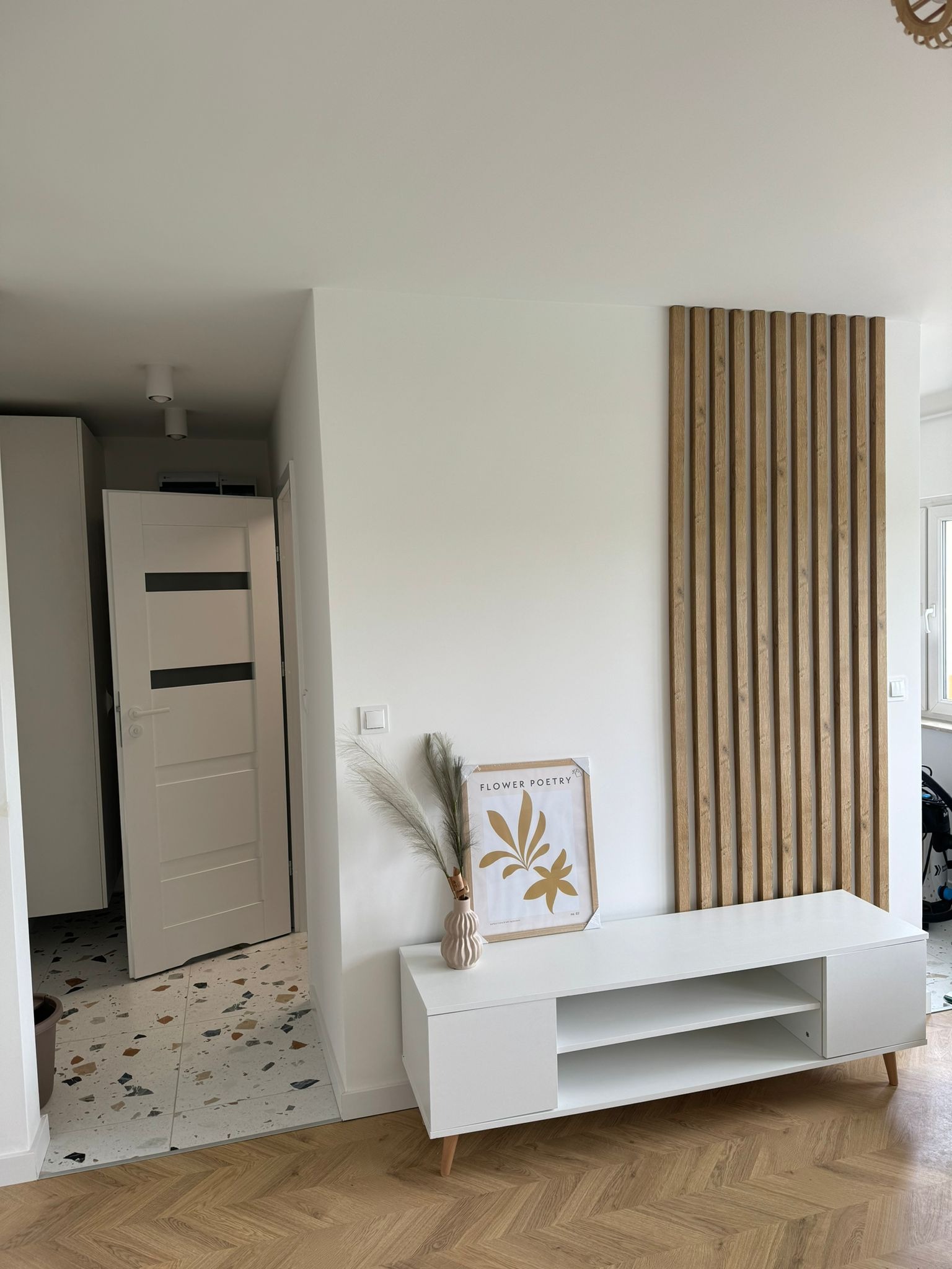 Nowoczesny przedpokój z białymi ścianami, drewnianą lamelą dekoracyjną, białą szafką RTV z dekoracjami, terazzo i parkietem w jodełkę.