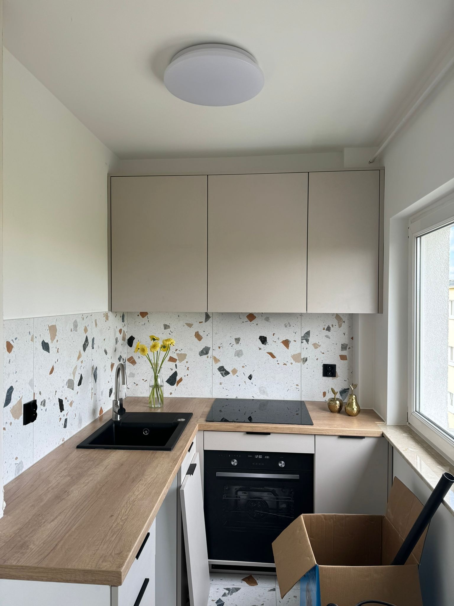 Nowoczesna kuchnia w trakcie wykańczania: beżowe szafki wiszące, blat imitujący drewno, czarny zlew i płyta indukcyjna, ściana z płytek w stylu lastryko, karton z akcesoriami na podłodze.