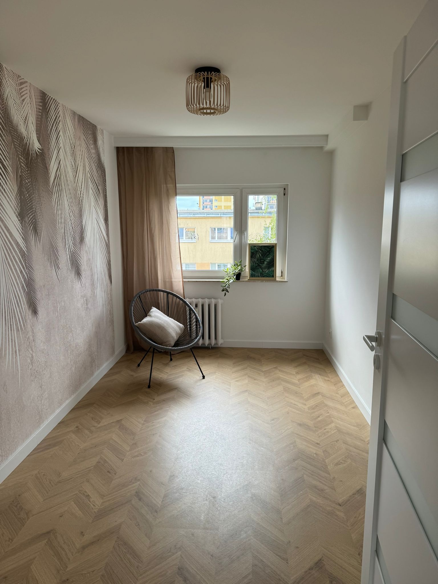 Pokój po remoncie: parkiet w jodełkę, ściana z tapetą w palmy, okno z roślinami, designerski fotel z poduszką i ażurowa lampa sufitowa.