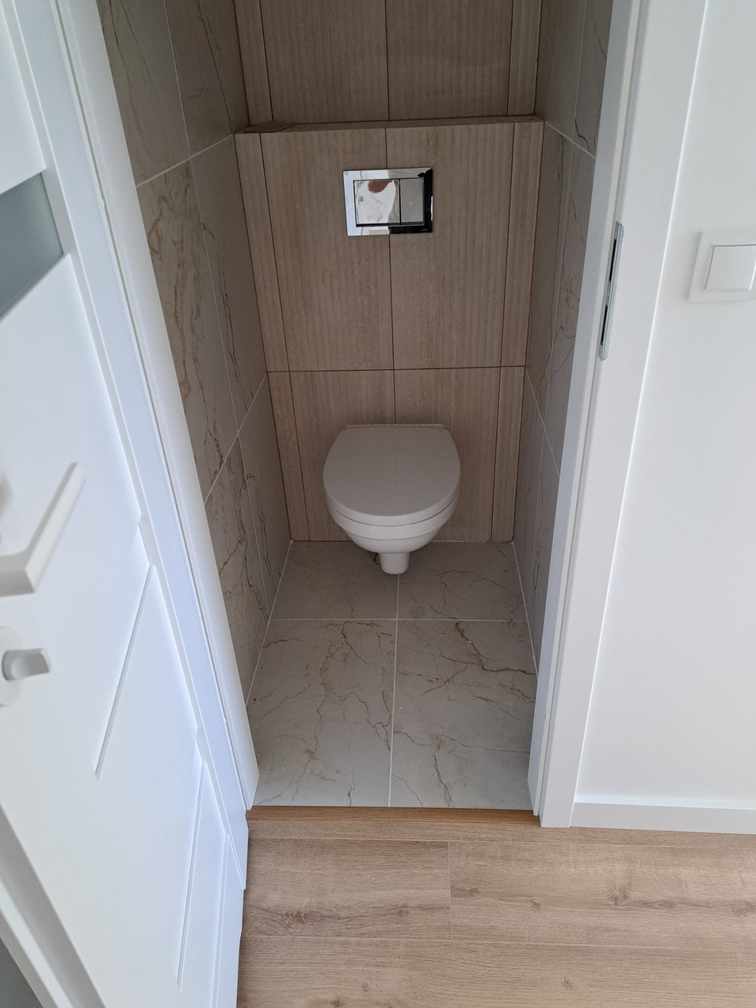 Mała toaleta wykończona płytkami imitującymi marmur, z podwieszaną miską WC i chromowanym przyciskiem spłukiwania, widok zza uchylonych białych drzwi.