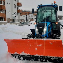 FHU ALEKSANDRA URBAN - Niebieski traktor z pomarańczowym pługiem odśnieża drogę na osiedlu mieszkaniowym. Zasypane śniegiem samochody w tle. Widoczny znak strefy ruchu.
