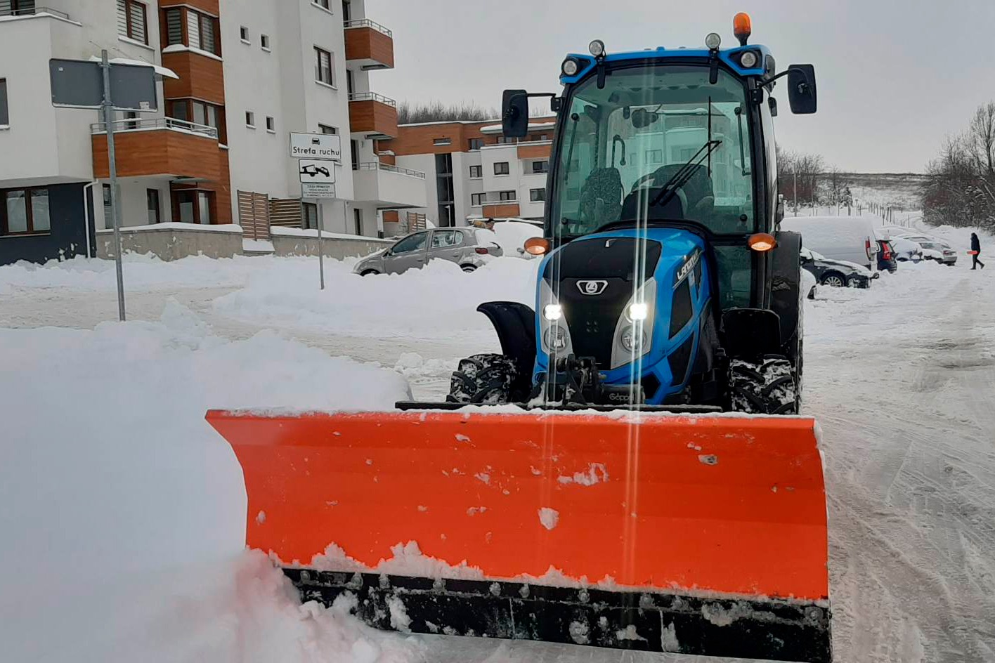 Niebieski traktor z pomarańczowym pługiem odśnieża drogę na osiedlu mieszkaniowym. Zasypane śniegiem samochody w tle. Widoczny znak strefy ruchu.