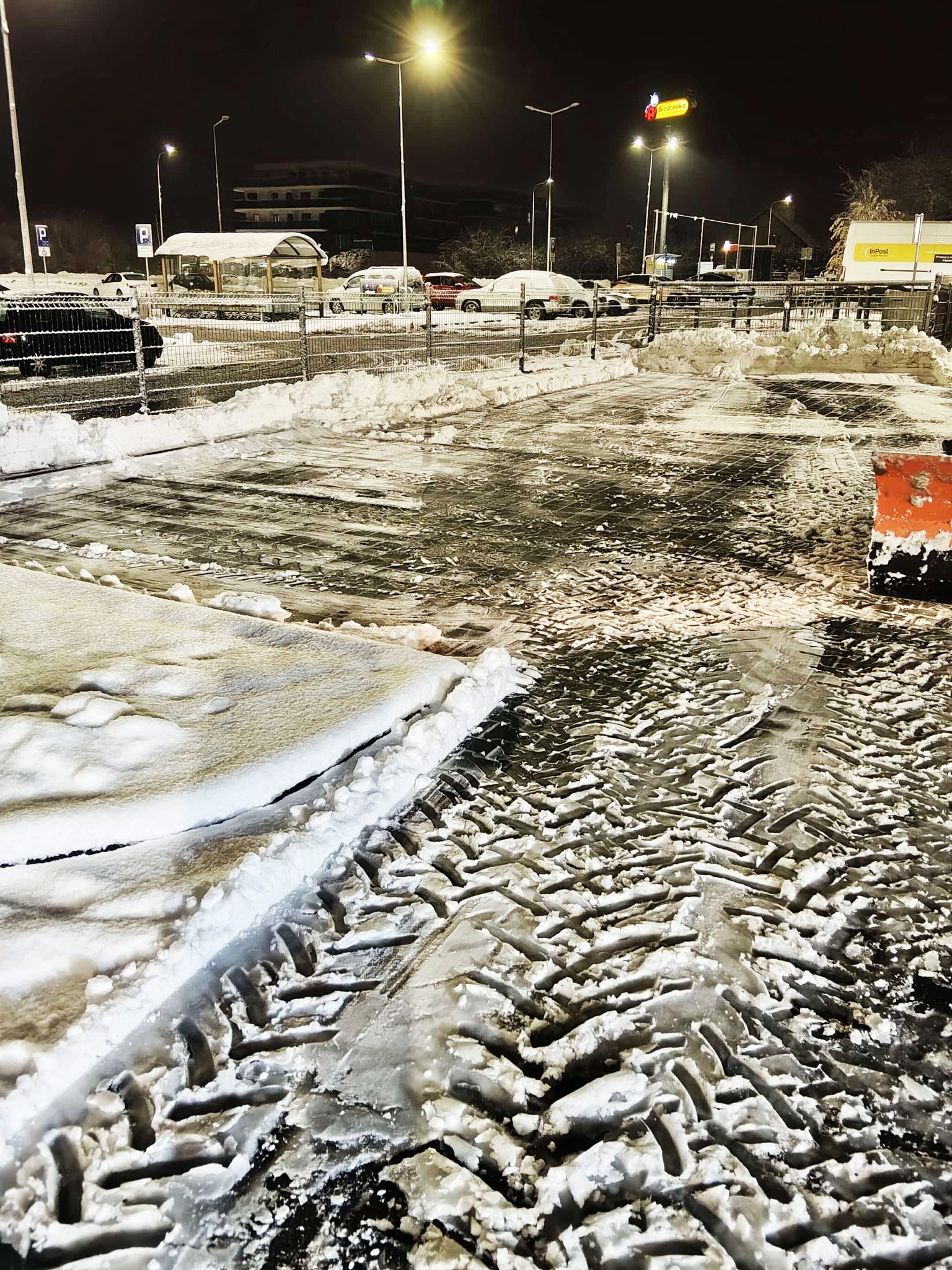 Odśnieżony parking nocą, widoczne ślady opon na mokrej nawierzchni, resztki śniegu przy krawężnikach i zaparkowane samochody w tle pod latarniami.