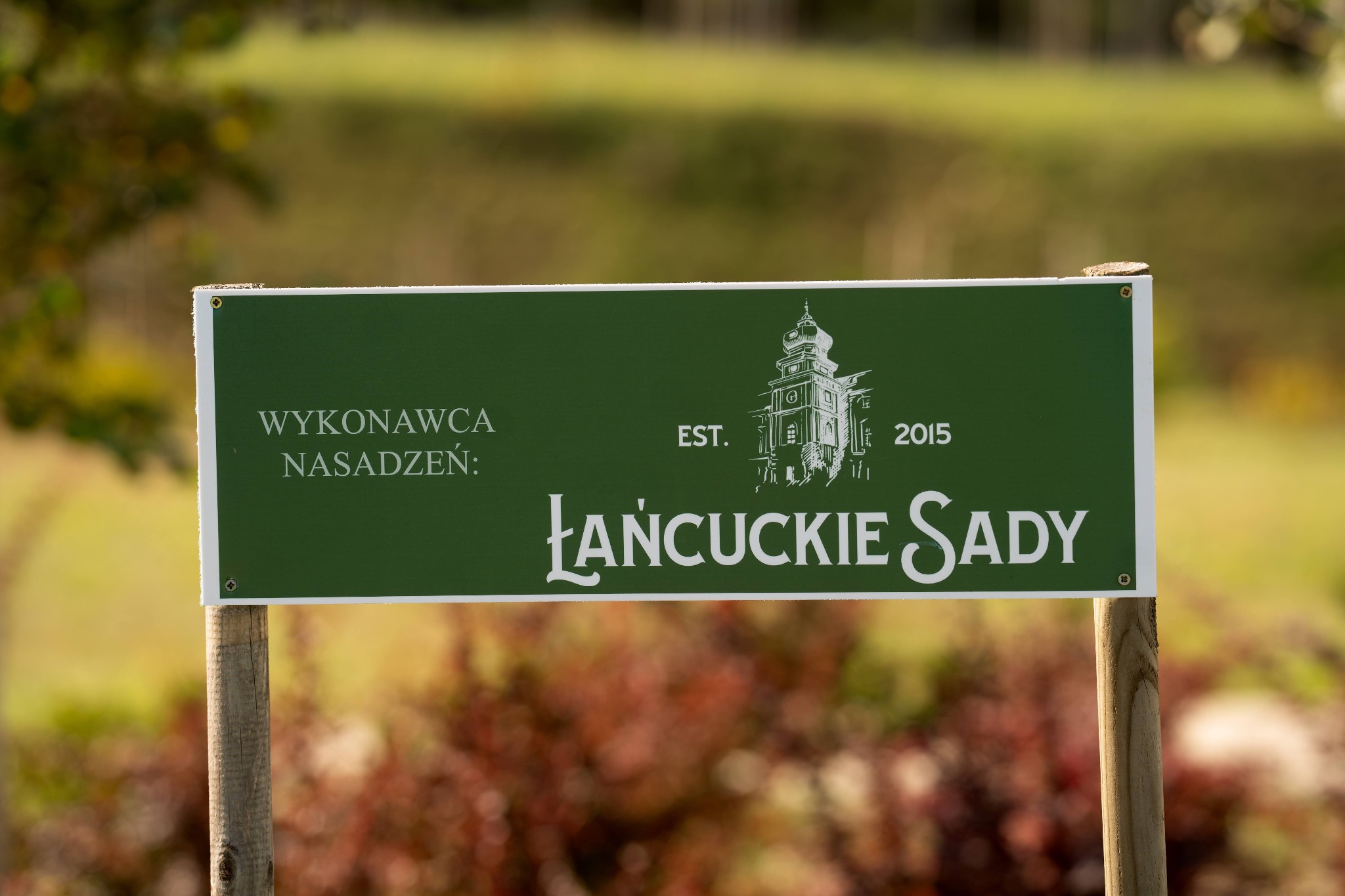 Tablica informacyjna z napisem 'Wykonawca nasadzeń: Łańcuckie Sady', rok założenia 2015, umieszczona na drewnianych słupkach na tle zieleni.
