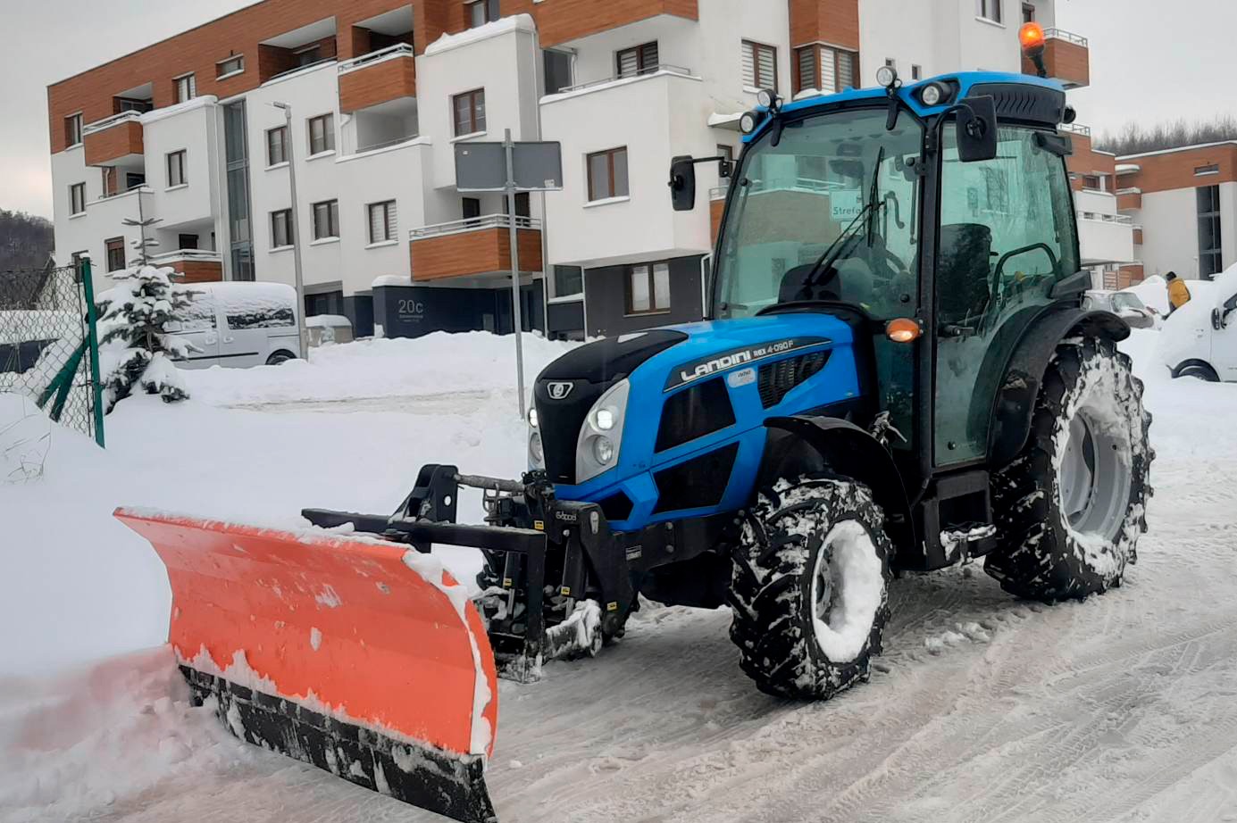 Niebieski traktor Landini z pomarańczowym pługiem odśnieżającym na tle ośnieżonego terenu i budynku mieszkalnego. Widoczne ślady opon na śniegu.