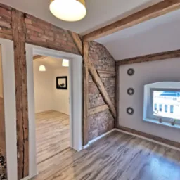 Wnętrze pokoju z drewnianą podłogą, ceglaną ścianą z drewnianymi belkami, oknem z widokiem na budynek i dekoracyjnymi lustrami na ścianie.