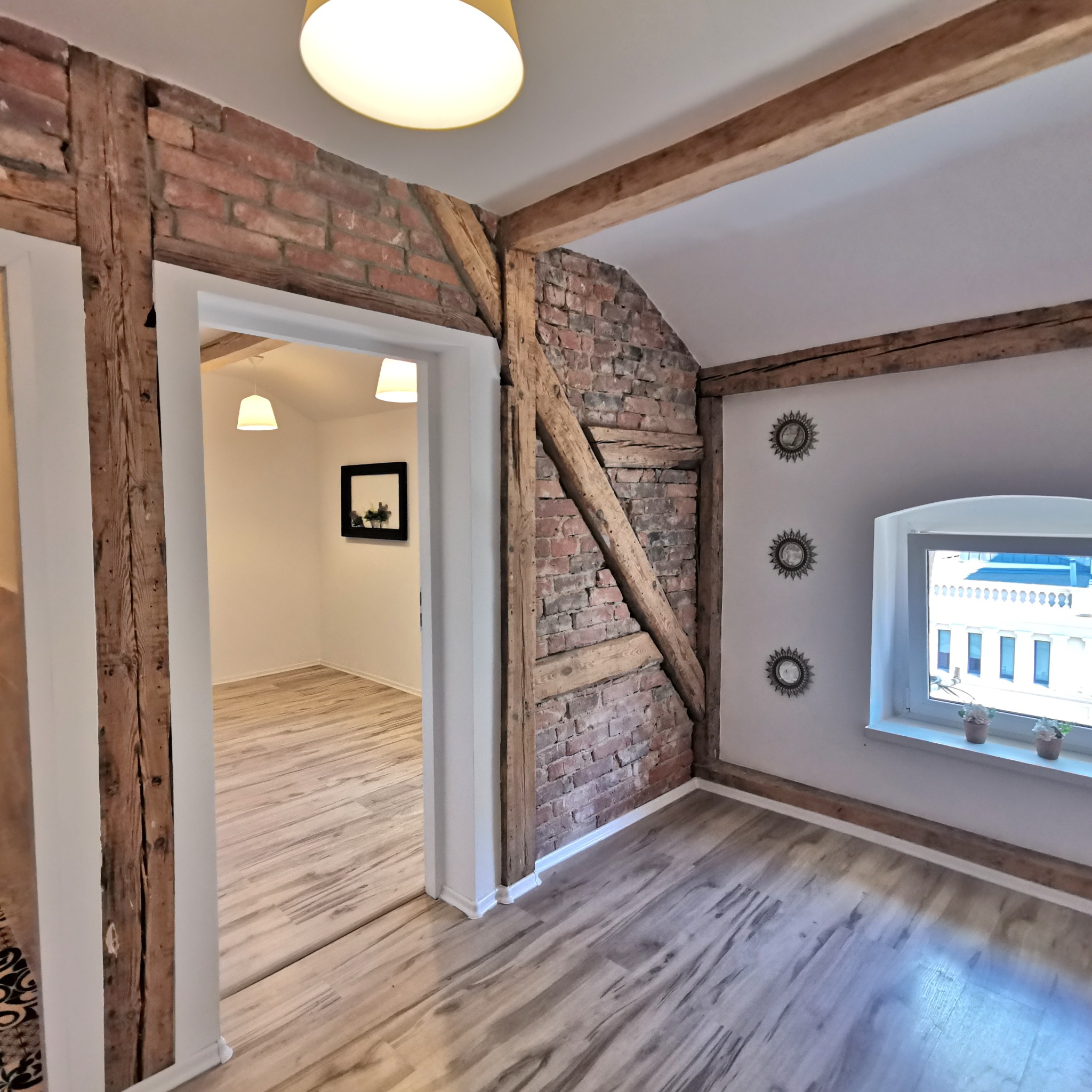 Wnętrze pokoju z drewnianą podłogą, ceglaną ścianą z drewnianymi belkami, oknem z widokiem na budynek i dekoracyjnymi lustrami na ścianie.