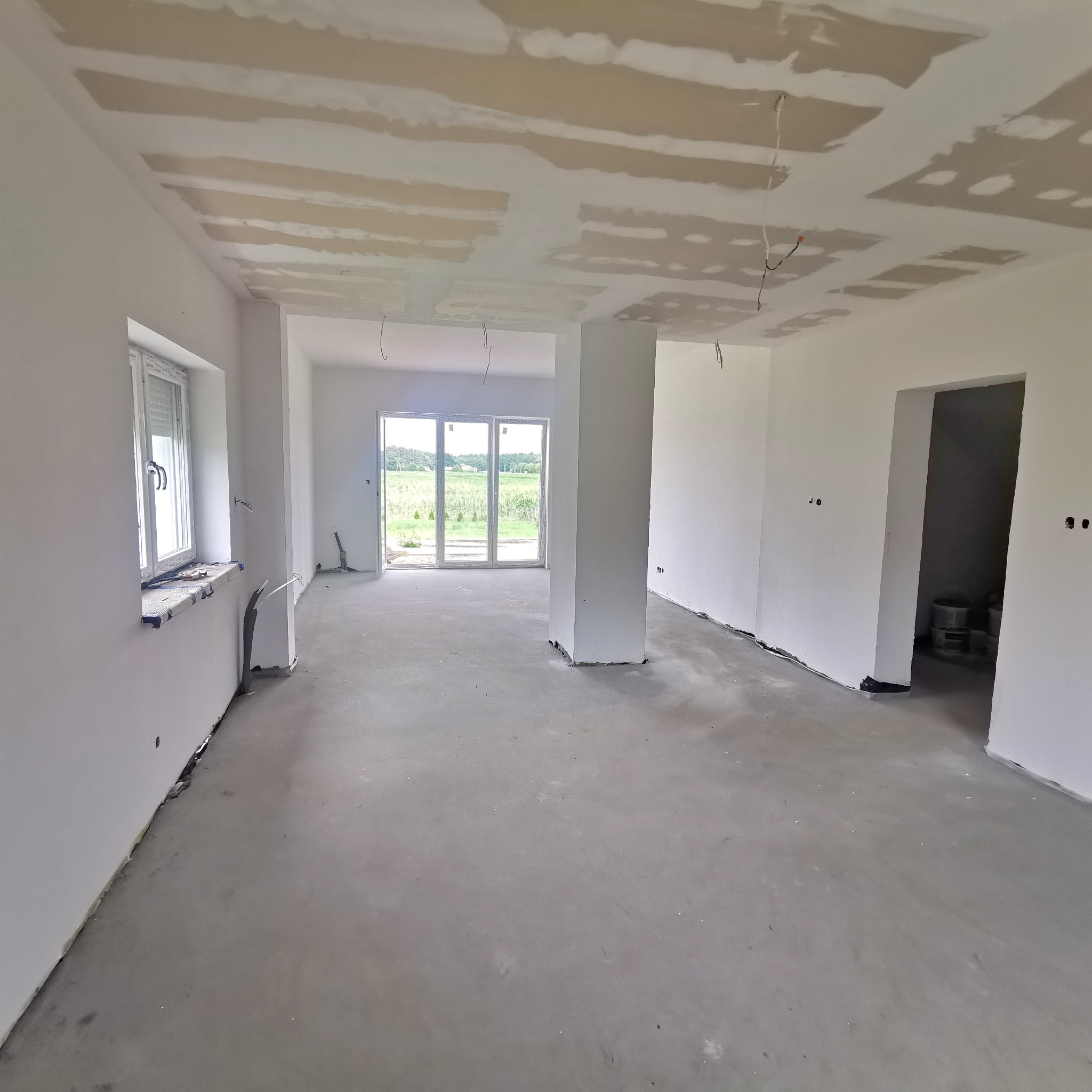 Wnętrze w trakcie prac wykończeniowych, widoczne ślady po szpachlowaniu na suficie, surowy beton na podłodze, ściany przygotowane do malowania, okno z roletami i widok na zewnątrz.