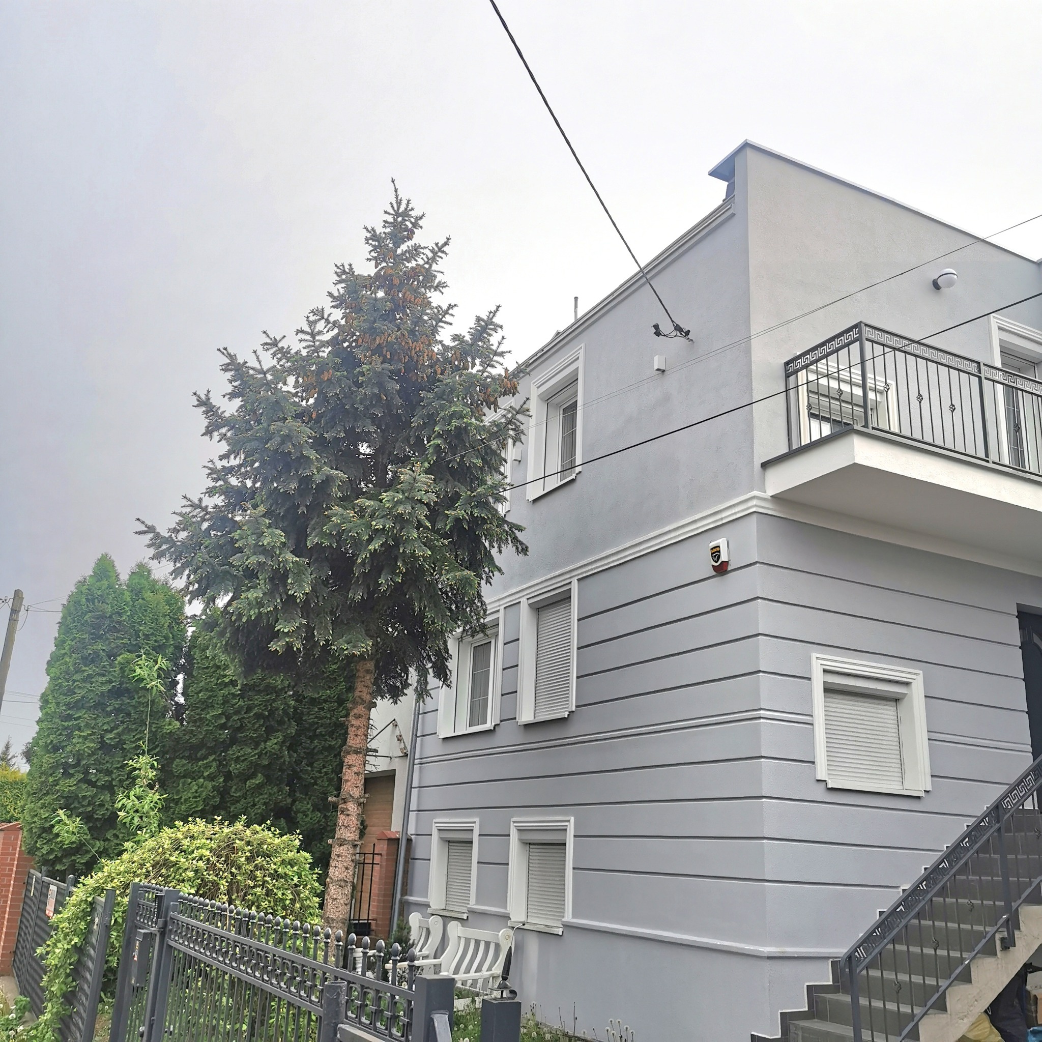Szary, dwupiętrowy dom z balkonem, widoczny zza ogrodzenia i drzew, z elewacją w poziome pasy i białymi obramowaniami okien.