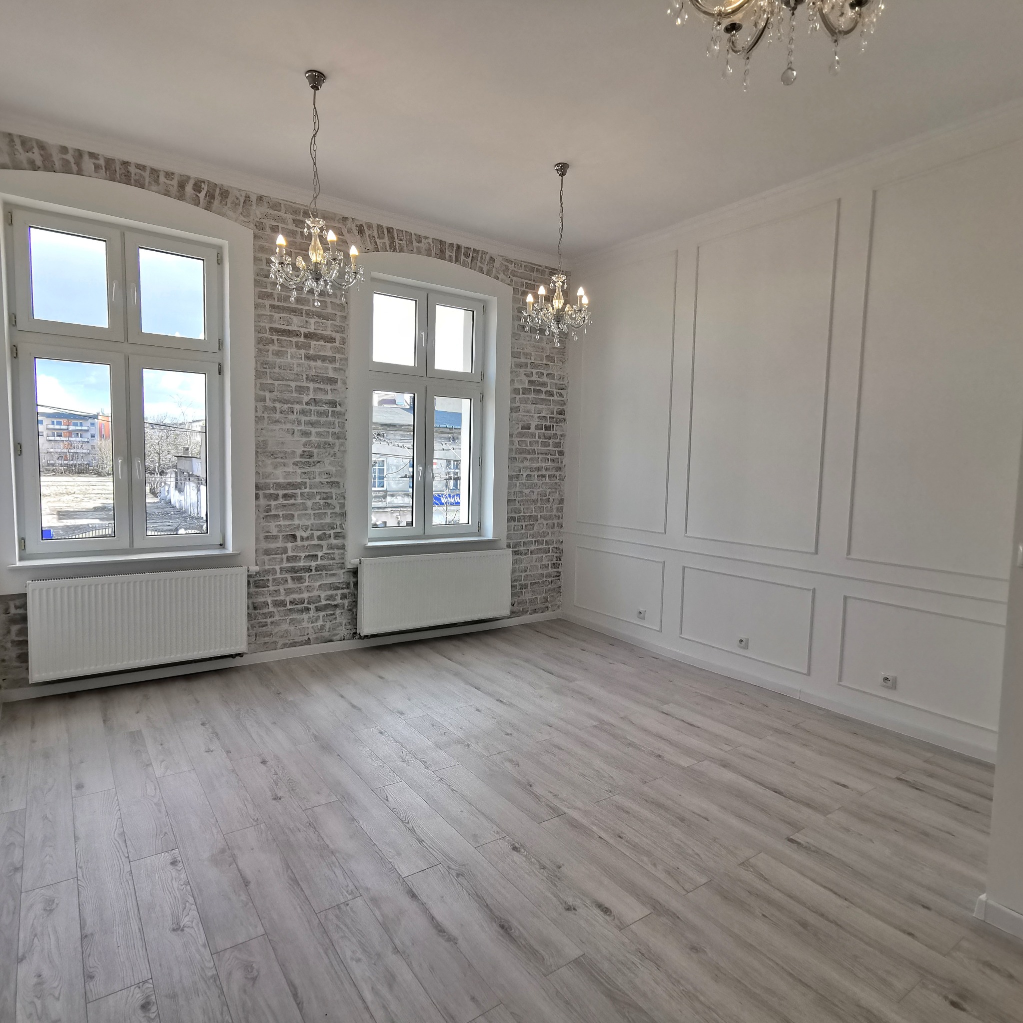 Puste, jasne pomieszczenie po remoncie z panelami podłogowymi, oknami w szarych, ceglanych wnękach, białymi grzejnikami i dekoracyjnymi listwami na ścianie. Wiszące, kryształowe żyrandole dodają...