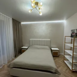 Nowoczesna sypialnia z łóżkiem, szafkami nocnymi i regałem, oświetlona lampą sufitową i listwą LED, z widocznymi zasłonami i parkietem w jodełkę.