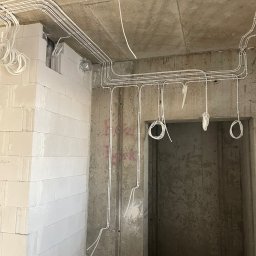 VOLTREND - Surowy stan: białe przewody elektryczne ułożone na suficie i ścianach w nowym budynku z betonu komórkowego, widoczne pętle przewodów zwisające z sufitu.