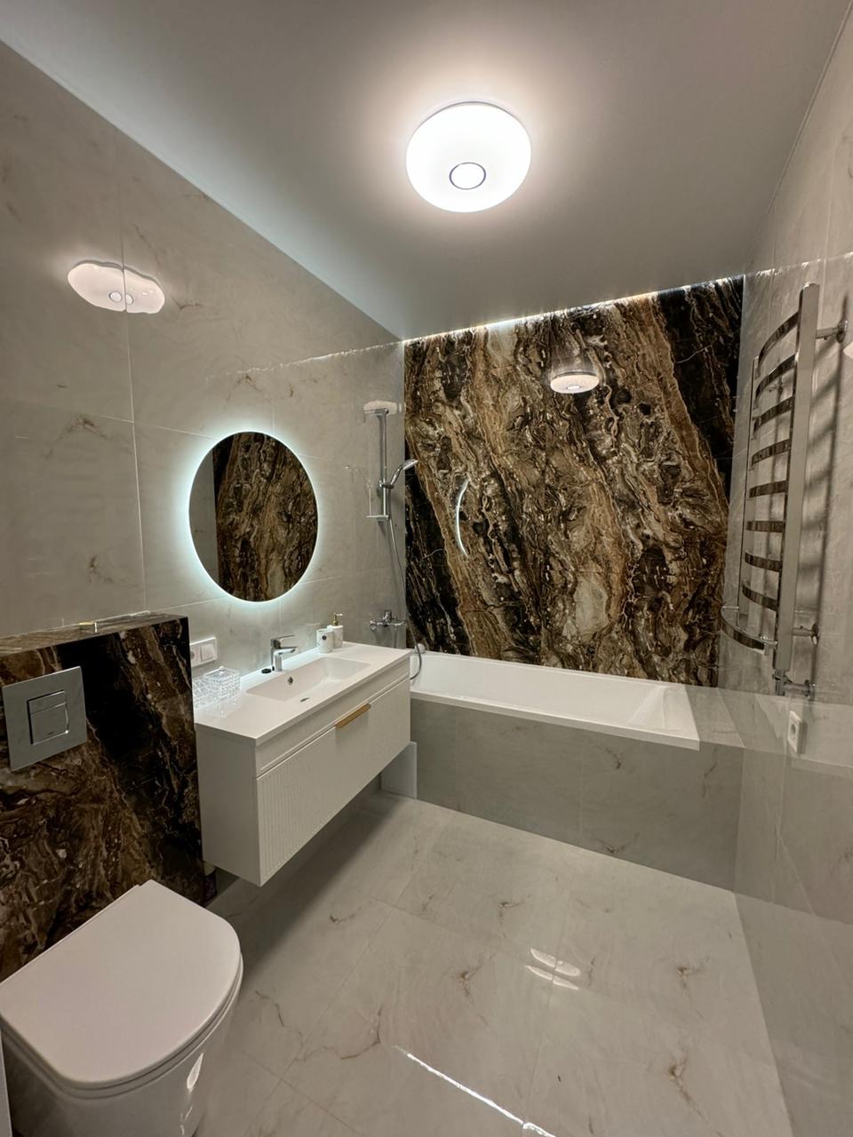 Nowoczesna łazienka z marmurowymi płytkami, okrągłym lustrem z podświetleniem LED i minimalistycznym oświetleniem sufitowym.