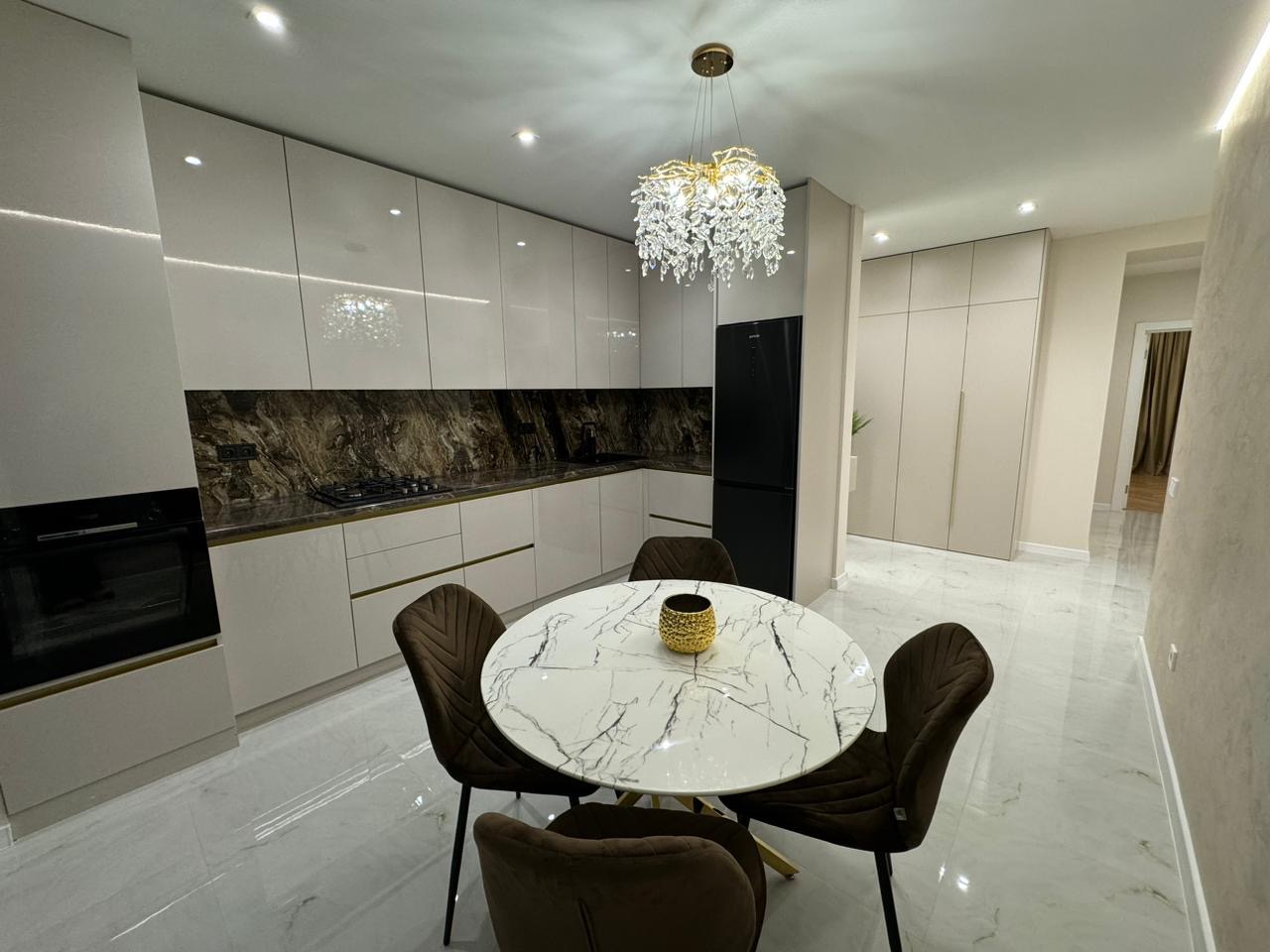 Nowoczesna kuchnia z białymi, połyskującymi szafkami, marmurowym blatem i backsplashiem, okrągłym stołem z marmurowym blatem oraz dekoracyjnym, kryształowym żyrandolem. Widoczne wbudowane sprzęty...