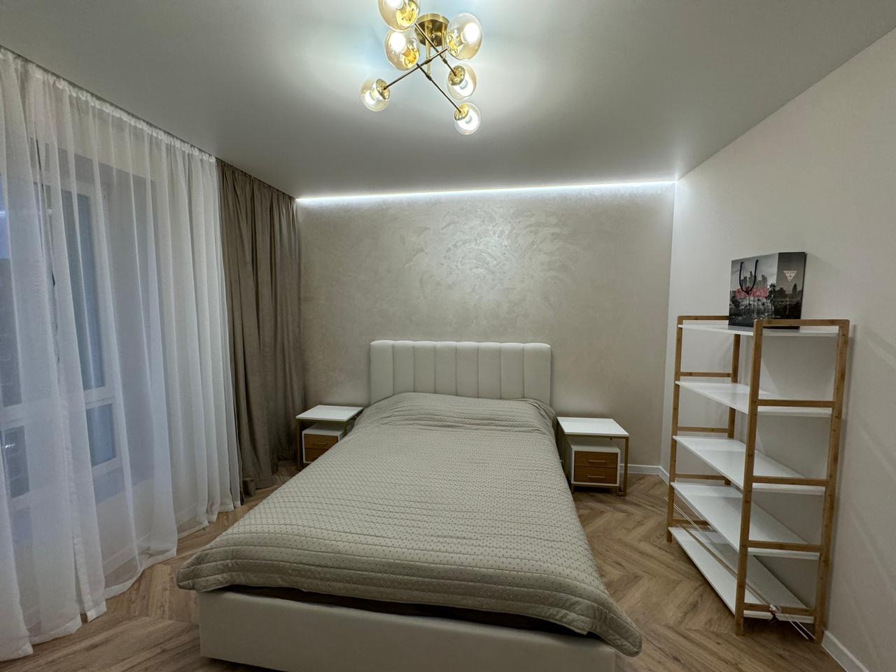 Nowoczesna sypialnia z łóżkiem, szafkami nocnymi i regałem, oświetlona lampą sufitową i listwą LED, z widocznymi zasłonami i parkietem w jodełkę.