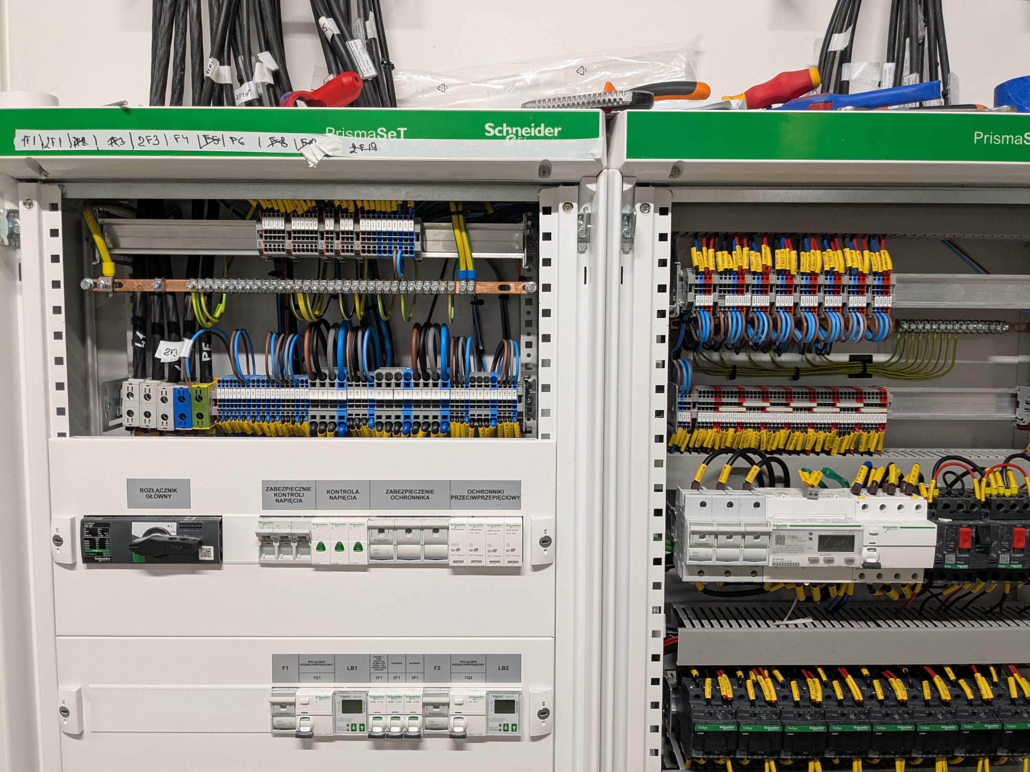 Rozdzielnica elektryczna Schneider PrismaSeT z widocznymi zabezpieczeniami, kontrolą napięcia, rozłącznikiem głównym oraz okablowaniem w kolorach: żółtym, niebieskim, czarnym i brązowym. Na górze...