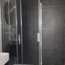 Chaus Remont Valerii Chaus - Nowoczesna kabina prysznicowa z deszczownicą w łazience wykończonej ciemnymi płytkami, widok z boku, z umywalką po lewej stronie. Minimalistyczny design.