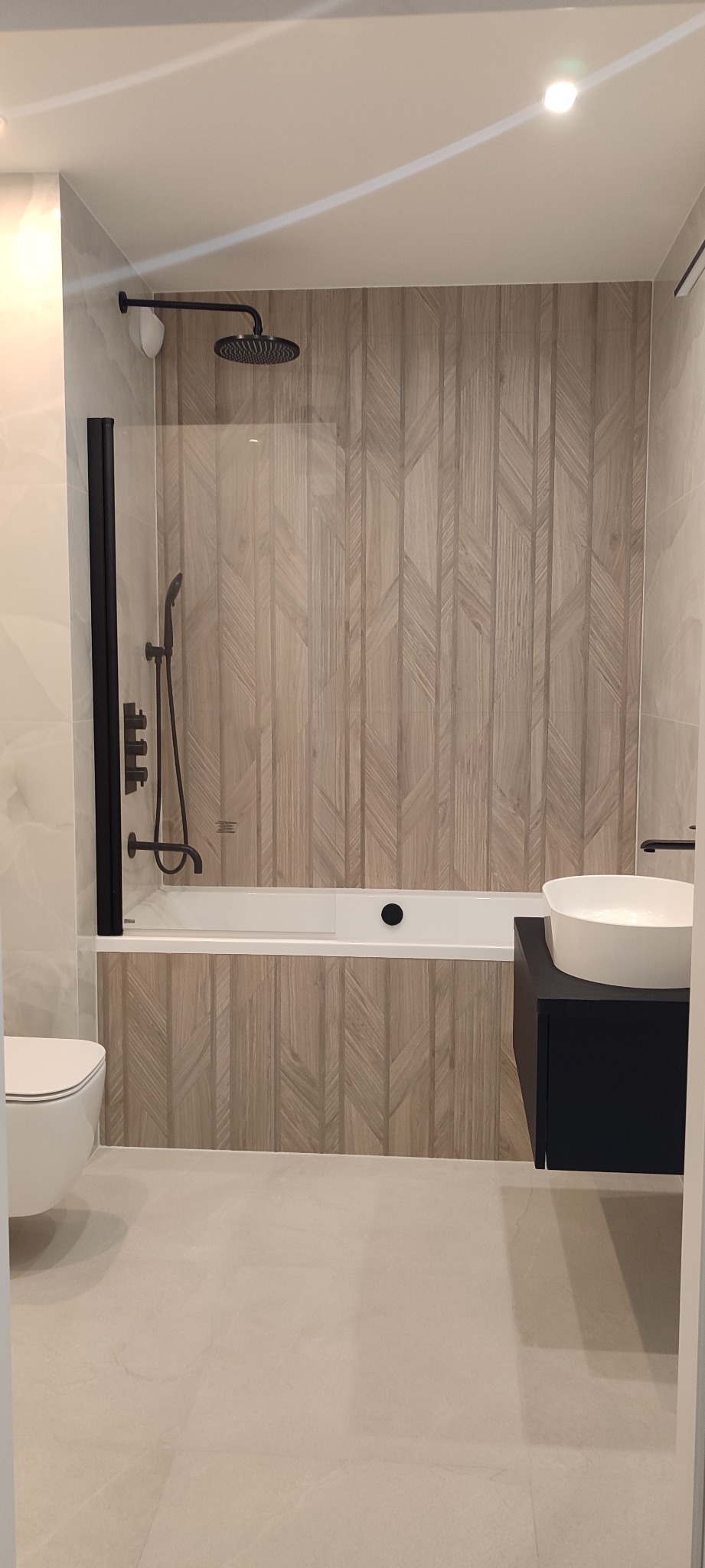 Nowoczesna łazienka po remoncie: drewnopodobne płytki chevron, czarna armatura, biała umywalka nablatowa i toaleta. Minimalistyczny design i jasna kolorystyka.