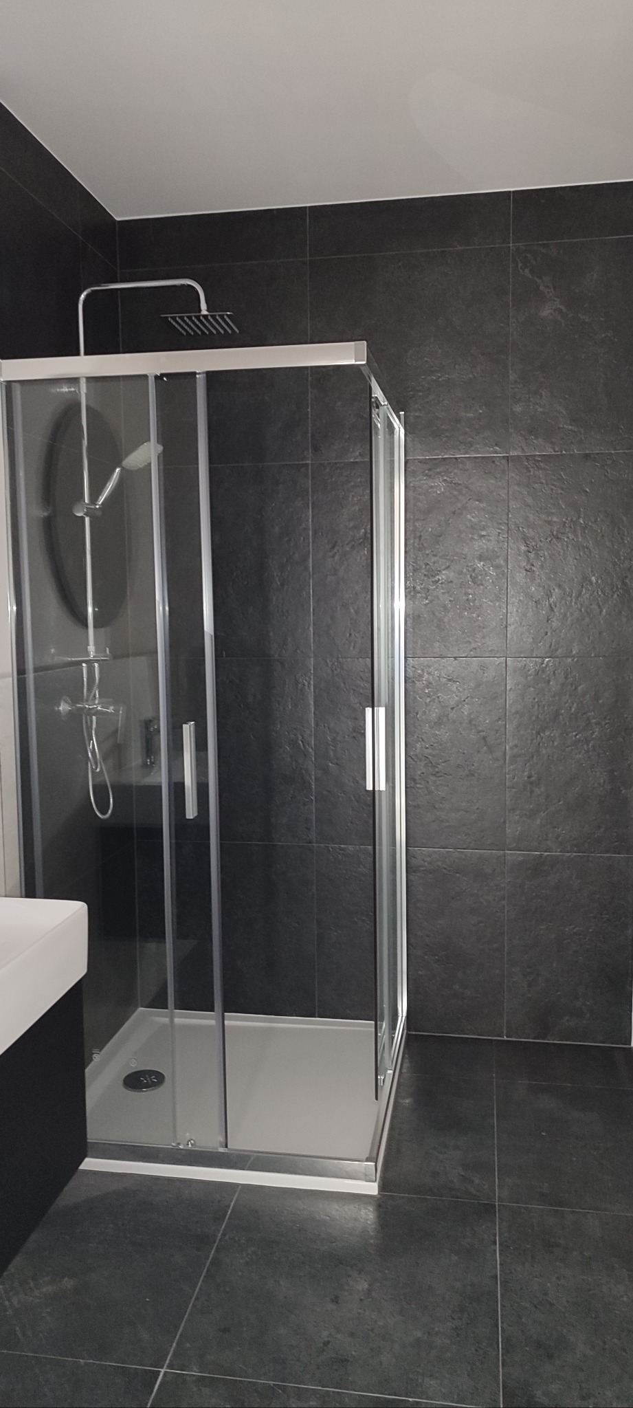 Nowoczesna kabina prysznicowa z deszczownicą w łazience wykończonej ciemnymi płytkami, widok z boku, z umywalką po lewej stronie. Minimalistyczny design.