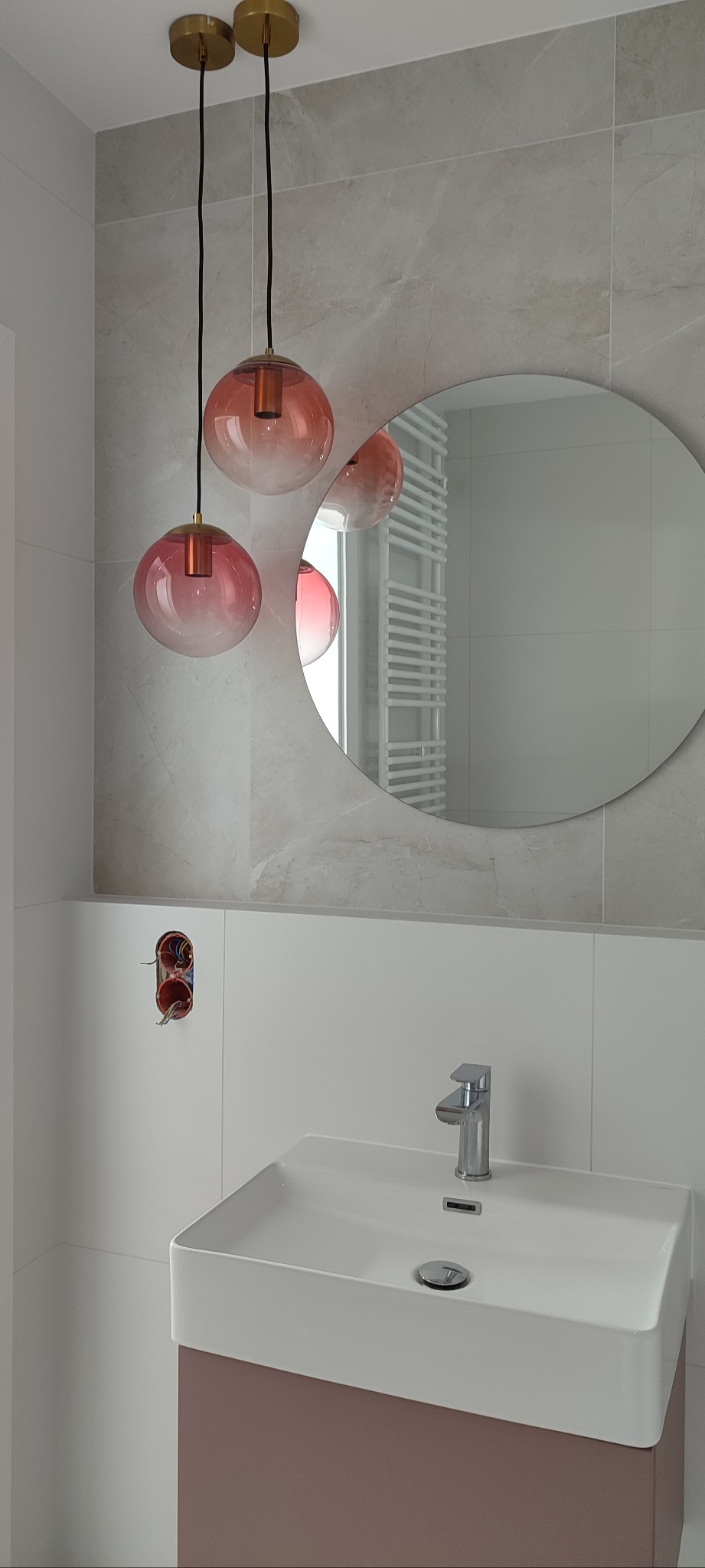 Nowoczesna łazienka z okrągłym lustrem, umywalką z szafką w kolorze pudrowego różu i trzema szklanymi lampami wiszącymi. Widoczne płytki imitujące marmur.