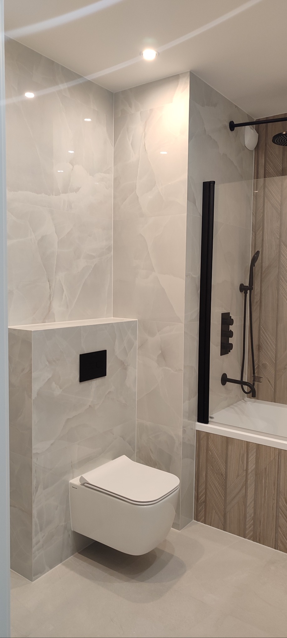 Elegancka łazienka z podwieszaną toaletą, płytkami imitującymi marmur i czarną armaturą. Kabina prysznicowa z drewnopodobnymi akcentami.
