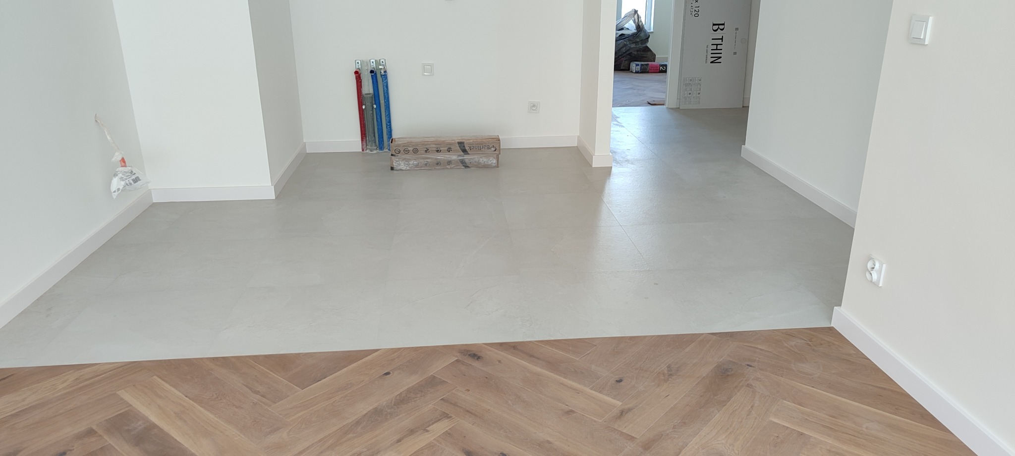 Jasne płytki podłogowe przechodzą w parkiet ułożony w jodełkę. Widoczne rury instalacyjne i kartony z panelami. Minimalistyczne, białe ściany i listwy przypodłogowe.