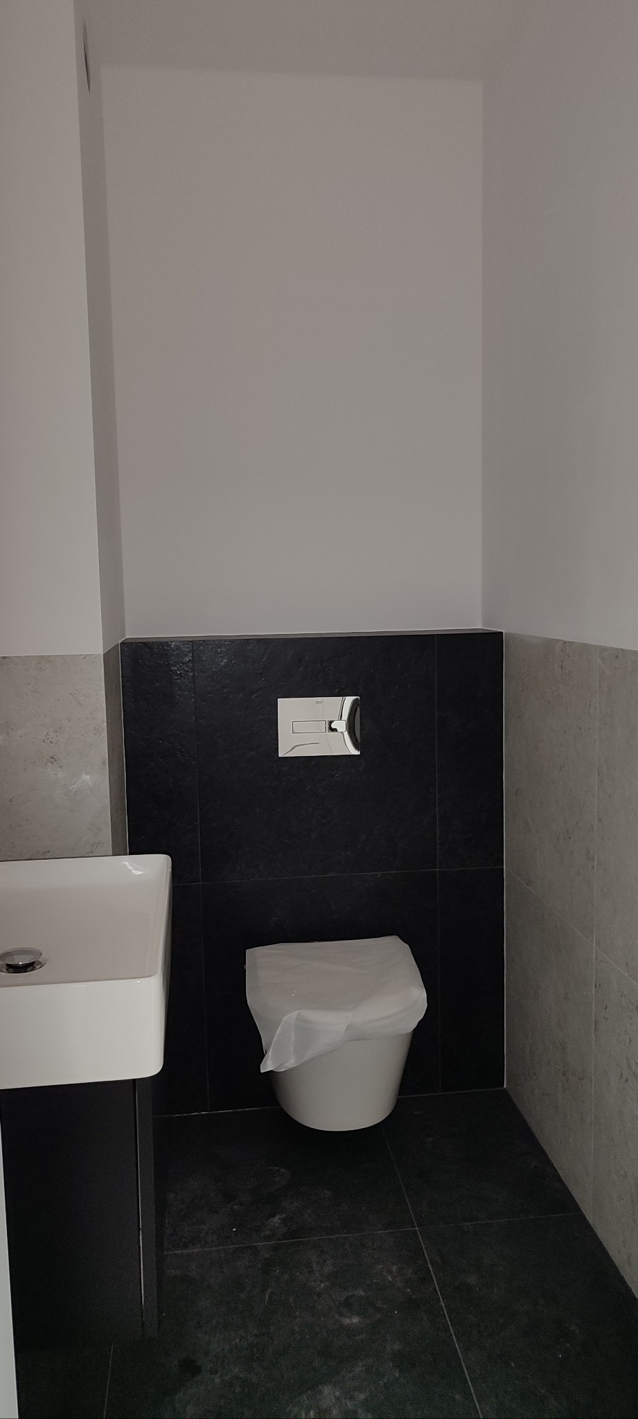 Nowoczesna łazienka: biała umywalka z czarną szafką, toaleta z przyciskiem spłukującym na tle czarnych i szarych płytek. Minimalistyczny design.