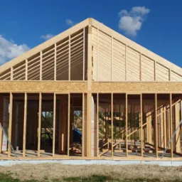 Konstrukcja domu w trakcie budowy z widocznym szkieletem drewnianym i elementami dachu na tle błękitnego nieba z chmurami.