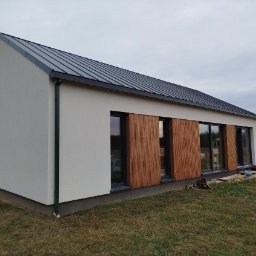 Nortegard Sp. z o.o. - Nowoczesny dom z jasną elewacją i drewnianymi panelami okiennymi, widok z zewnątrz w pochmurny dzień. Minimalistyczny design z ciemnym dachem.