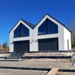 Nortegard Sp. z o.o. - Biały, nowoczesny dom z dwuspadowym dachem i dużymi oknami, z szarymi roletami garażowymi. Uwagę przyciągają schody prowadzące do wejścia i częściowo wykończony teren wokół.