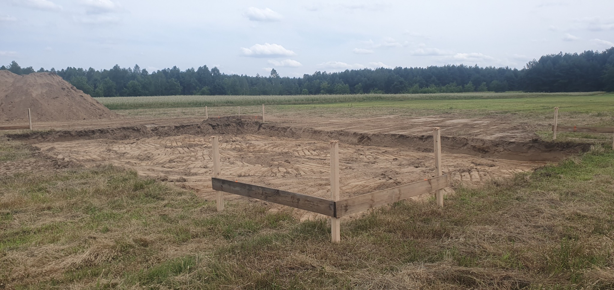 Przygotowanie terenu pod budowę, widoczne wykopane fundamenty, drewniane pale, ziemia i stos ziemi w tle, krajobraz wiejski.