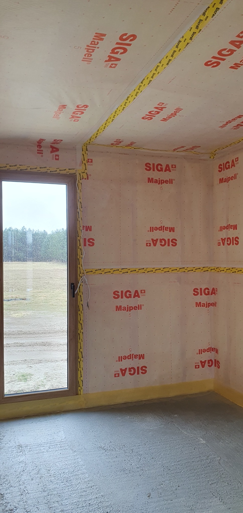 Wnętrze w trakcie budowy, ściany wyłożone membraną paroizolacyjną z nadrukiem Siga Majpell, uszczelnione żółtą taśmą, widoczne okno z widokiem na las i wylana betonowa posadzka.