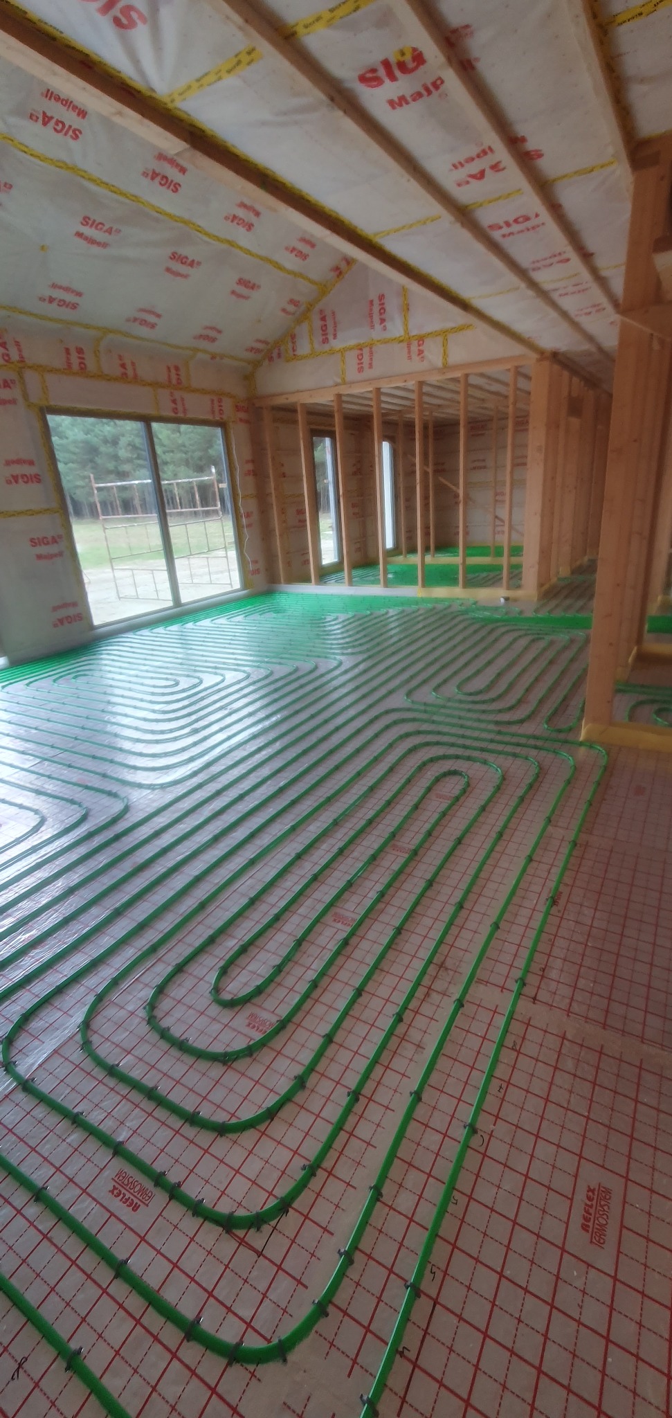 Wnętrze w stanie surowym otwartym z widoczną instalacją ogrzewania podłogowego z zielonych rurek ułożonych na izolacji podłogowej w regularnych pętlach, ściany w konstrukcji szkieletowej...