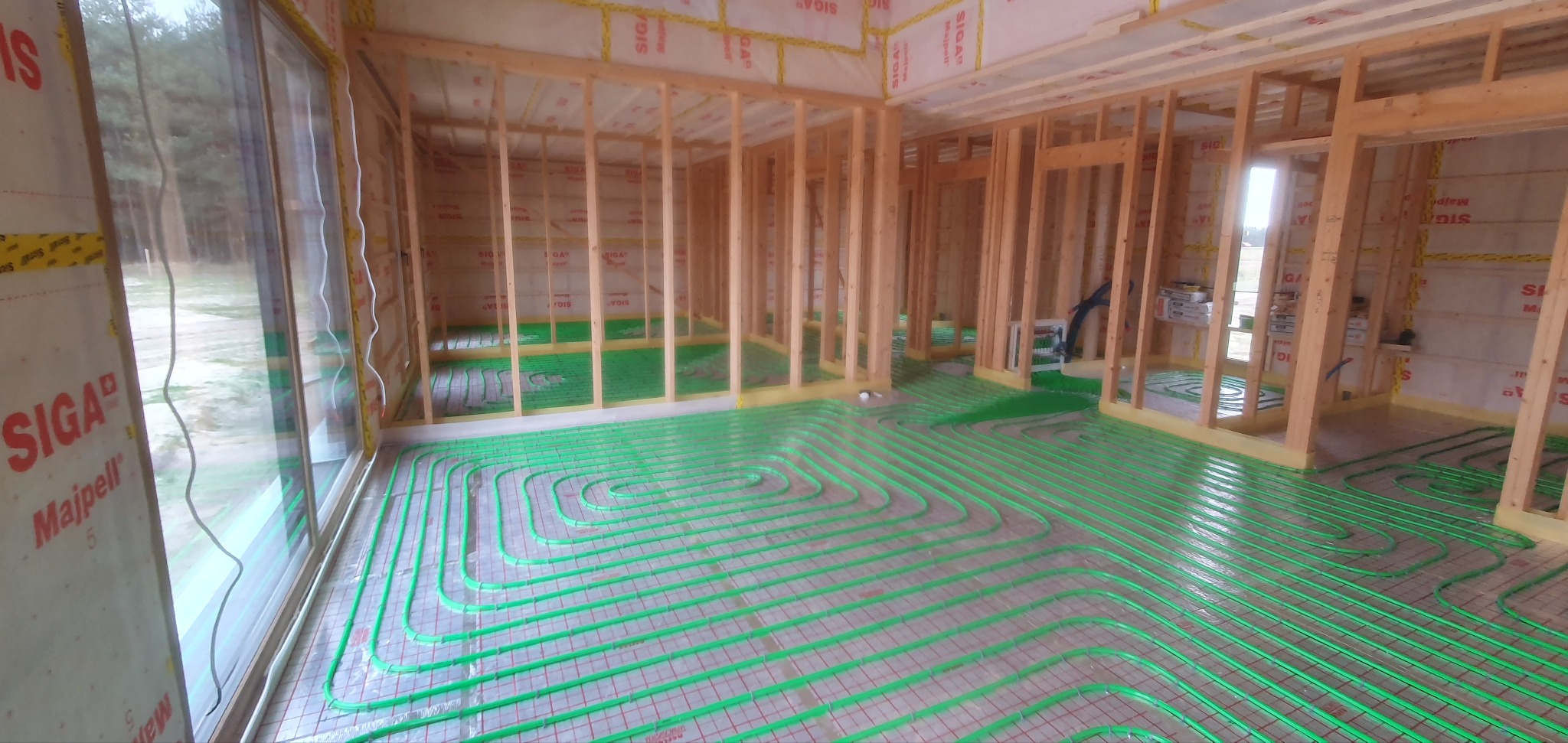 Wnętrze domu szkieletowego w trakcie budowy z widoczną instalacją ogrzewania podłogowego z zielonych rur ułożonych na folii izolacyjnej i drewnianą konstrukcją ścian.