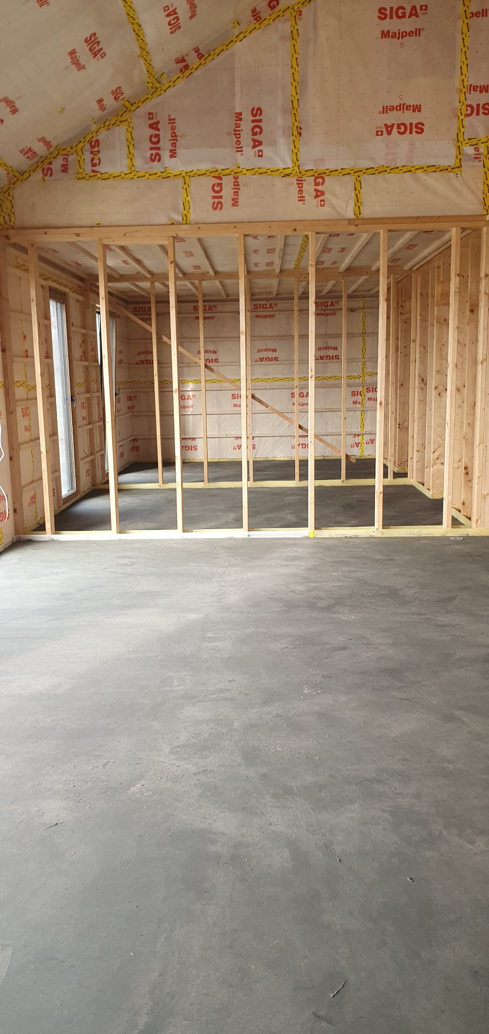 Wnętrze w trakcie budowy: surowy beton na podłodze, drewniana konstrukcja ścian z widoczną izolacją i folią paroizolacyjną z nadrukami producenta, okno po lewej stronie.