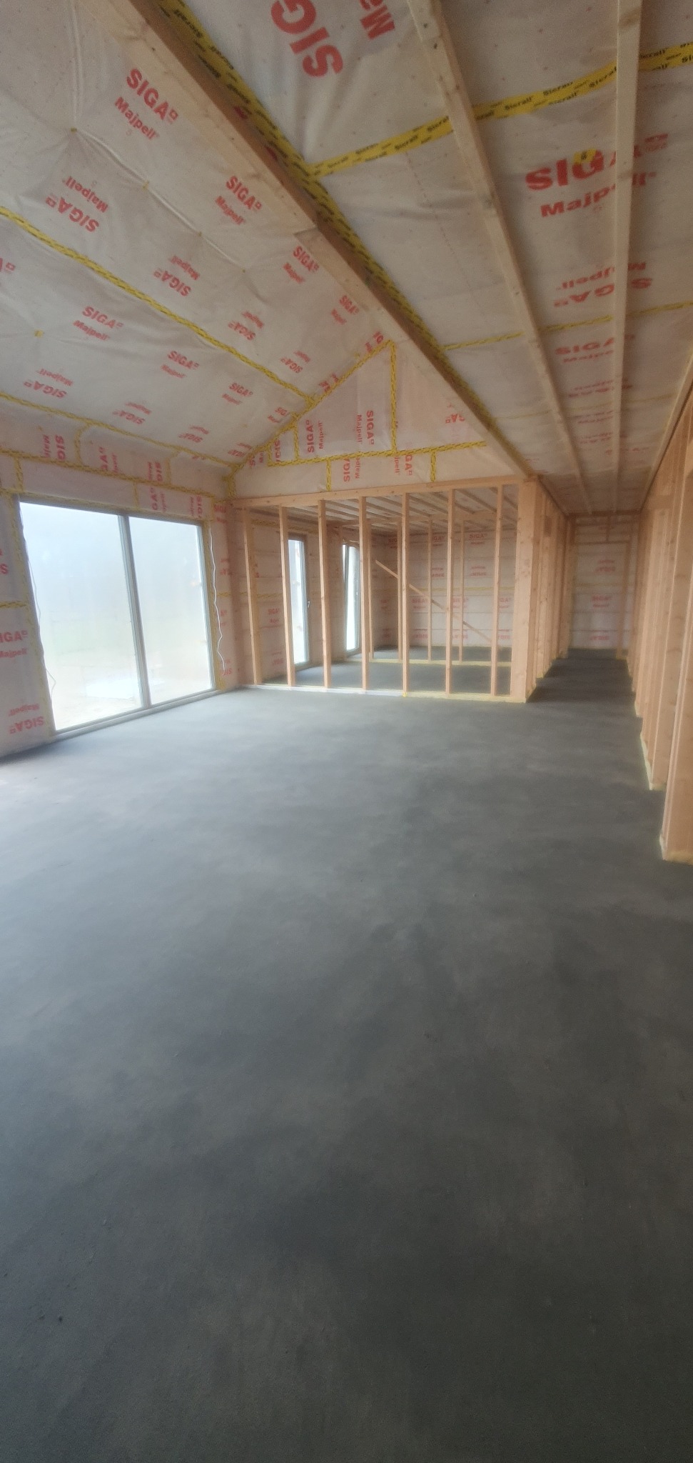 Wnętrze surowego domu w trakcie budowy, widoczne drewniane ramy ścian działowych, duże okna i szara wylewka na podłodze, z folią izolacyjną na skośnym dachu.