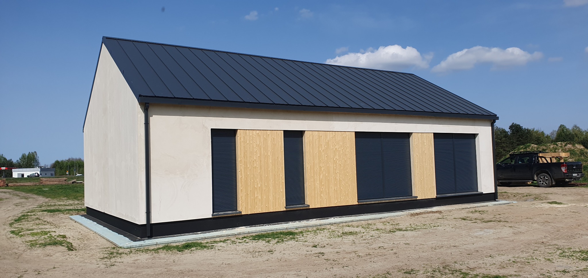 Gotowy dom z jasną elewacją, drewnianymi panelami i grafitowymi roletami zewnętrznymi, z grafitowym dachem na tle błękitnego nieba i zaparkowanego pickupa z drewnem w tle.