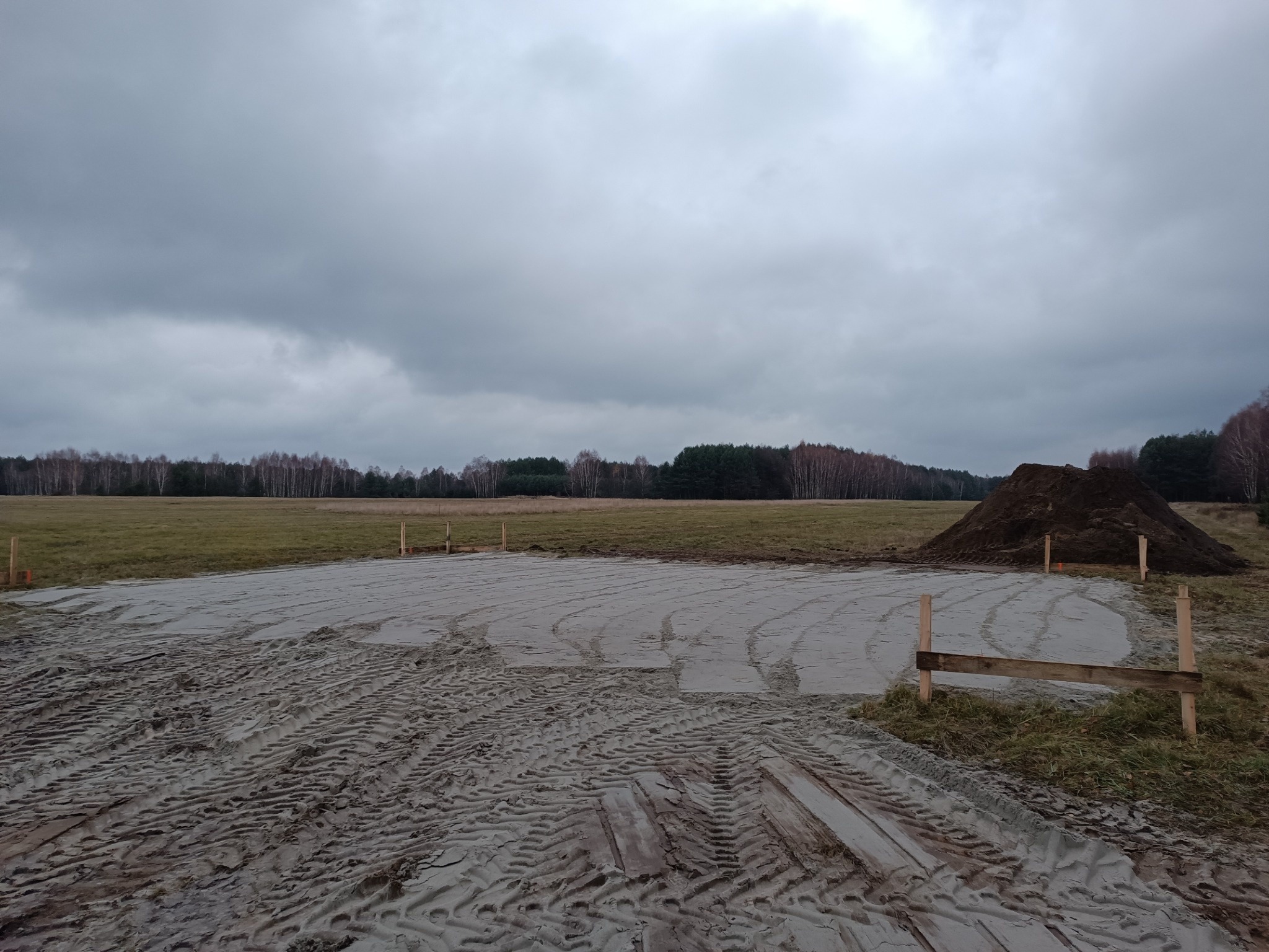 Przygotowany teren pod budowę z wytyczonym obrysem fundamentów, widoczny nasyp ziemi i ślady opon maszyn budowlanych, pochmurne niebo.