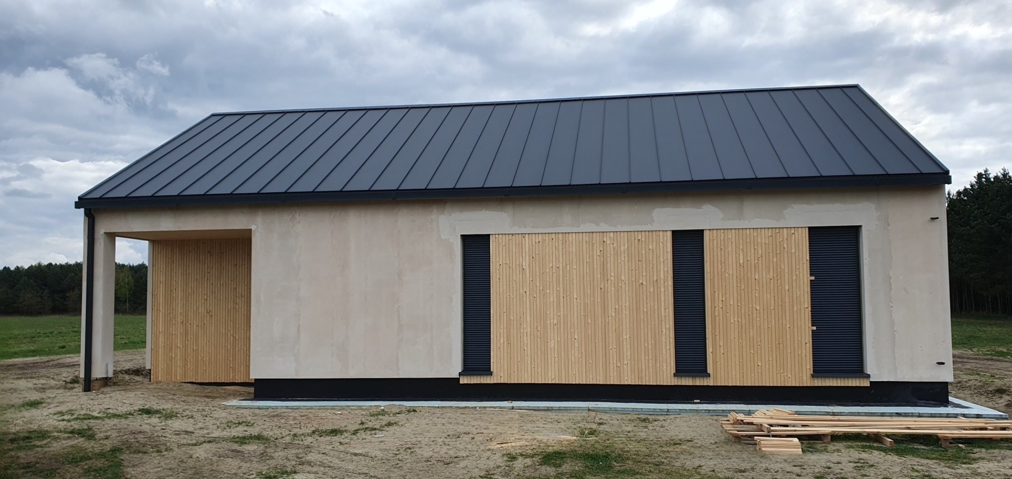 Niedokończony budynek z szarą blachodachówką, elewacją w kolorze piasku i drewnianą fasadą z czarnymi roletami, widoczne deski na ziemi.