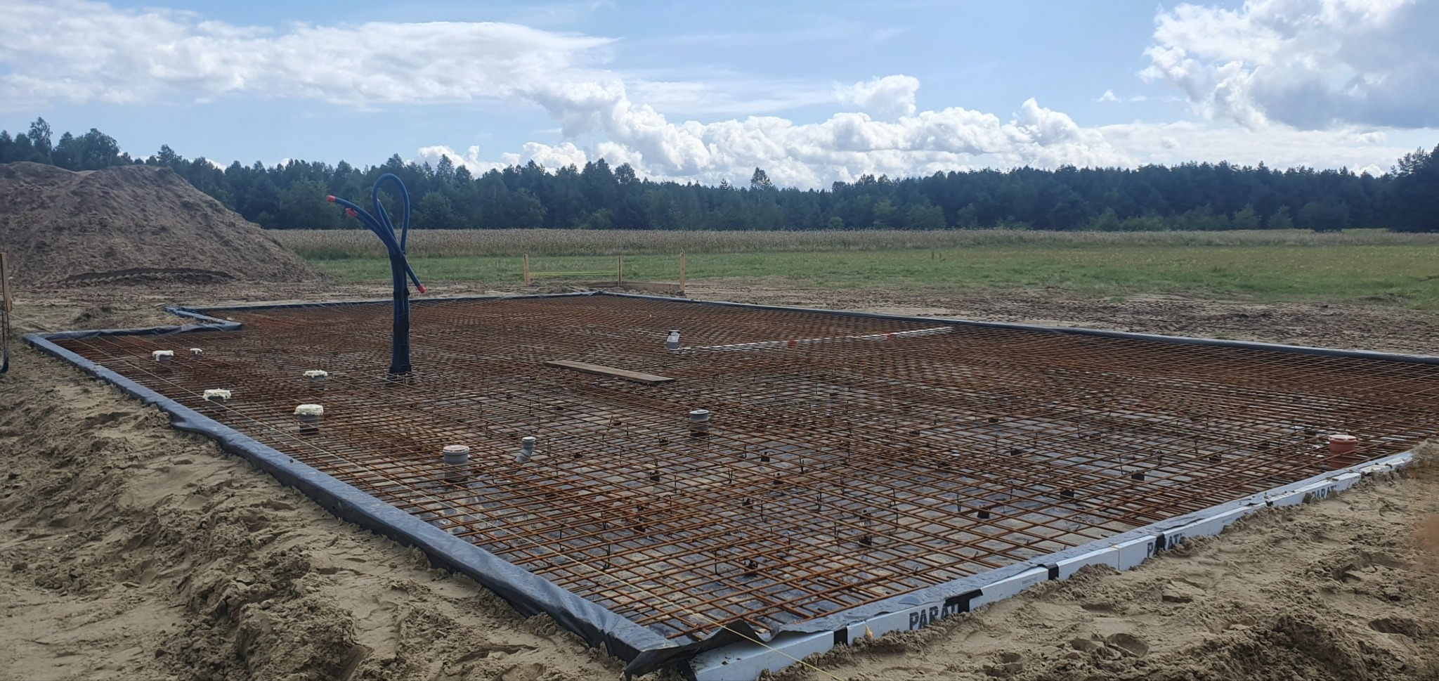 Przygotowanie zbrojenia płyty fundamentowej pod budowę domu na piaszczystym terenie; widoczne rury instalacyjne, siatka zbrojeniowa i izolacja obwodowa.