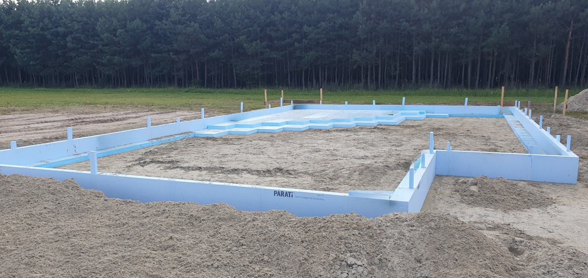 Fundament z izolacją termiczną z jasnoniebieskiego styropianu, przygotowany pod budowę domu, widok na las w tle.