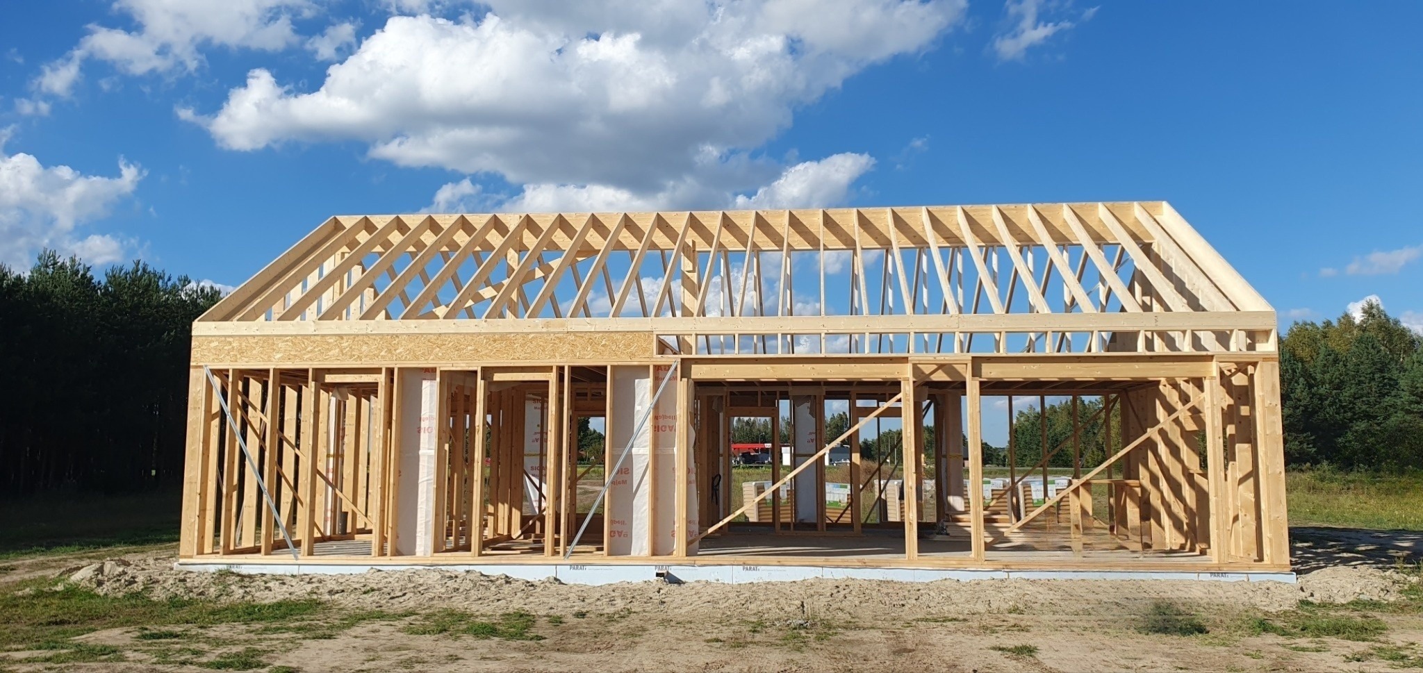 Wznoszenie konstrukcji domu z drewnianych belek na fundamencie, widoczne elementy ścian i dachu, na zewnątrz krajobraz z zielenią i błękitnym niebem z chmurami.