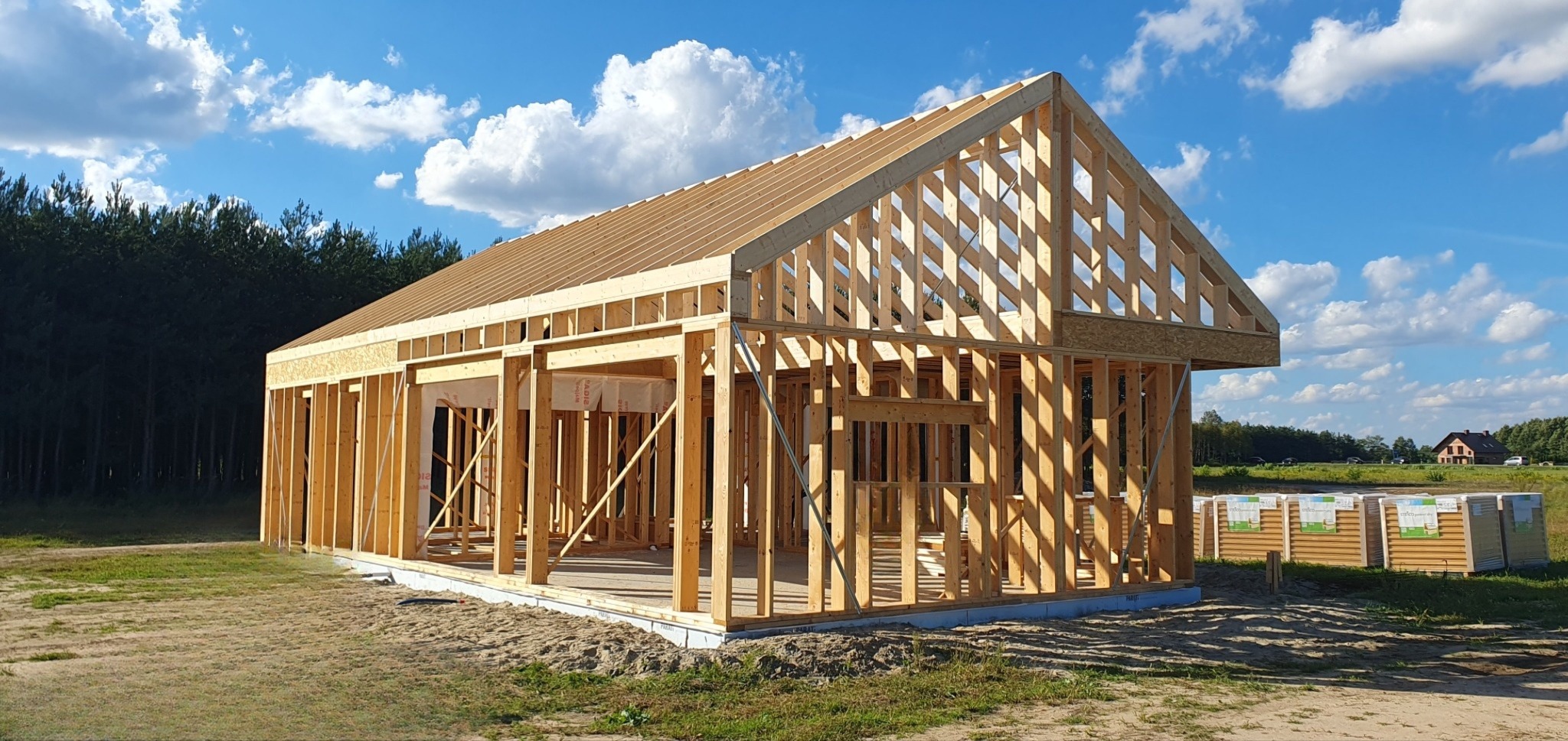 Konstrukcja drewniana domu w trakcie budowy, widoczne elementy szkieletu, więźba dachowa oraz składowane panele na zewnątrz, w tle las i błękitne niebo z chmurami.