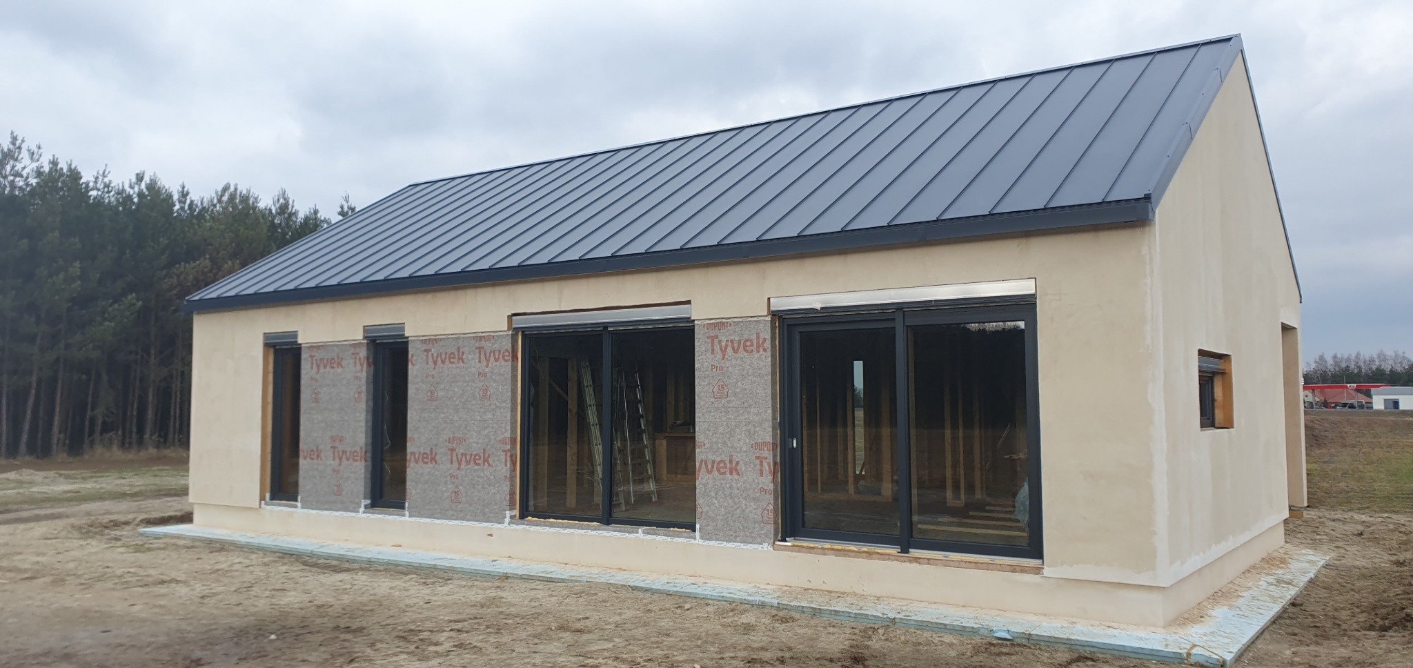 Niedokończony parterowy budynek z ciemnym dachem z blachy trapezowej, okna zabezpieczone folią Tyvek, widoczne rusztowania wewnątrz, na zewnątrz piasek.
