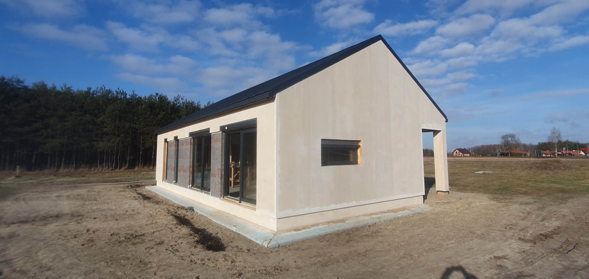 Surowy stan domu z czarnym dachem, z oknami zabezpieczonymi folią, na działce z widocznym lasem w tle.