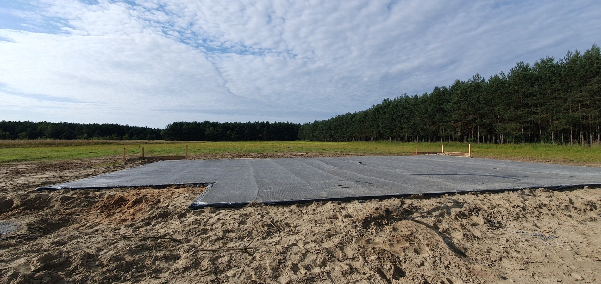 Przygotowany teren pod budowę z rozłożoną geowłókniną i wyznaczonym obrysem fundamentów, widok na rozległe pole i las w tle.