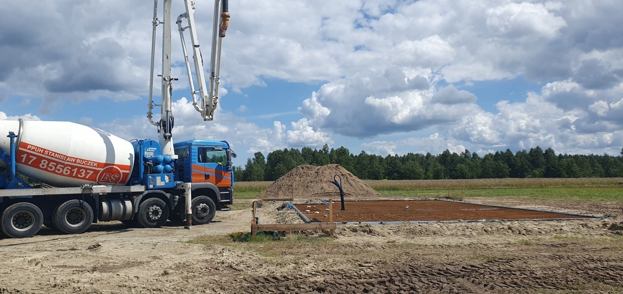 Betoniarka z wysięgnikiem na placu budowy, przygotowana do wylewania fundamentów pod budynek, widoczne zbrojenie i szalunek.