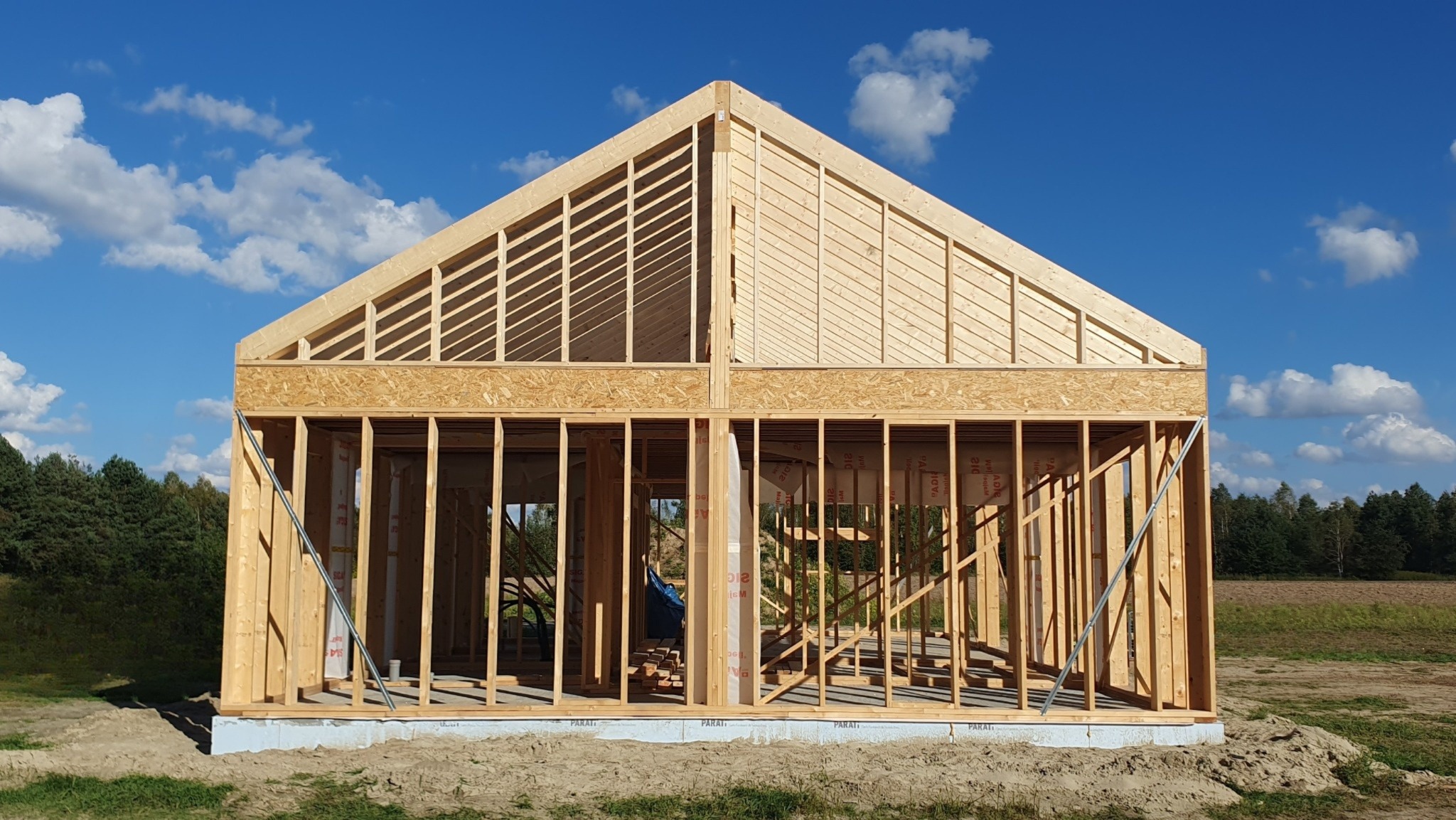 Konstrukcja domu w trakcie budowy z widocznym szkieletem drewnianym i elementami dachu na tle błękitnego nieba z chmurami.
