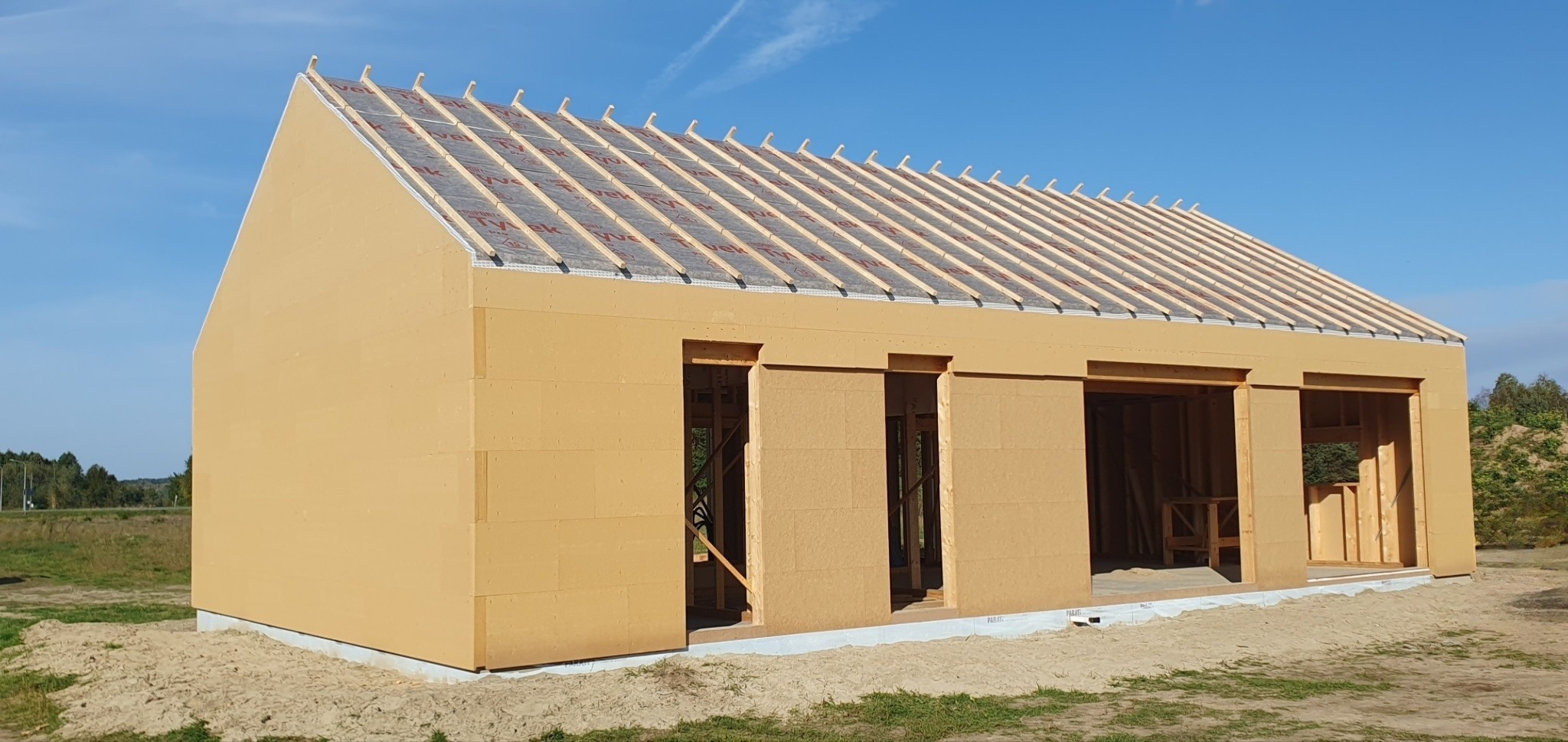 Niedokończony, parterowy budynek z płyt warstwowych o jasnobrązowej barwie, z widoczną konstrukcją dachu i otworami okiennymi oraz drzwiowymi, na piaszczystym terenie z fragmentami zieleni.