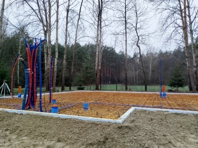 Przygotowanie fundamentów pod budowę domu: izolacja, instalacje wodno-kanalizacyjne, zbrojenie. Widok na las w tle, pochmurny dzień.